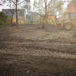 Żółta koparko-ładowarka JCB 432E na placu budowy, częściowo niwelującym teren pod zabudowę, widoczne ślady opon na ziemi i drzewa w tle.