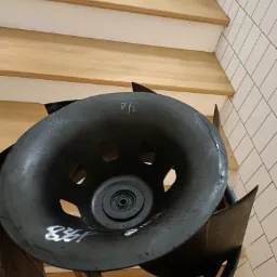 Czarny wentylator z widocznymi śladami mycia i numerem '834' naniesionym białą farbą, umieszczony na tle drewnianych schodów i białych kafelków.