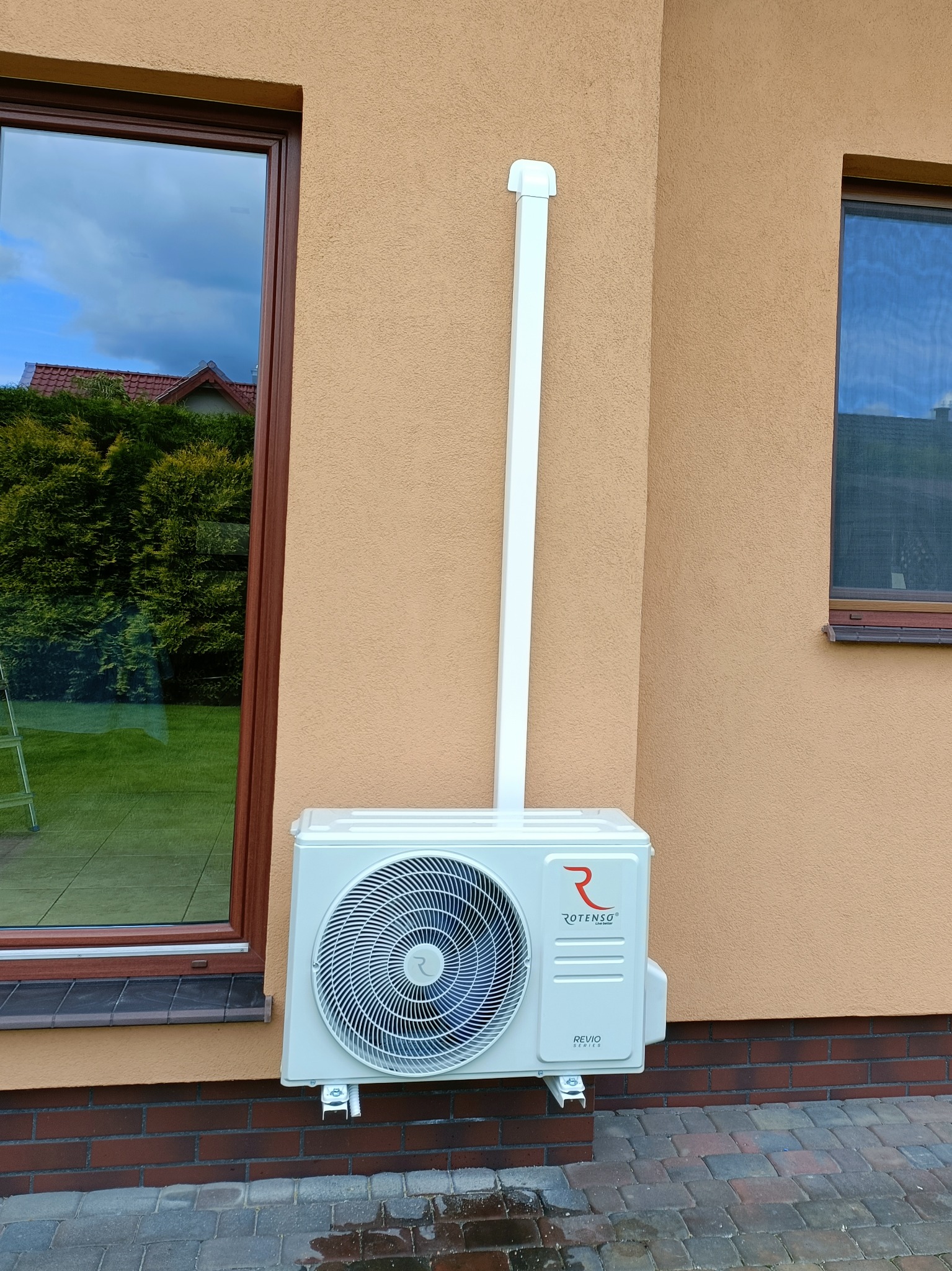 Zewnętrzna jednostka klimatyzacji Rotenso Revio 3,5 kW zamontowana na elewacji budynku, widoczna rura odprowadzająca skropliny, okno z odbiciem zieleni w tle.