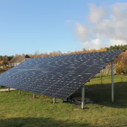 Instalacja fotowoltaiczna na trawniku przed domem jednorodzinnym w jesiennej scenerii, widoczne panele słoneczne, metalowa konstrukcja wsporcza, tło stanowią drzewa z żółtymi liśćmi i błękitne...