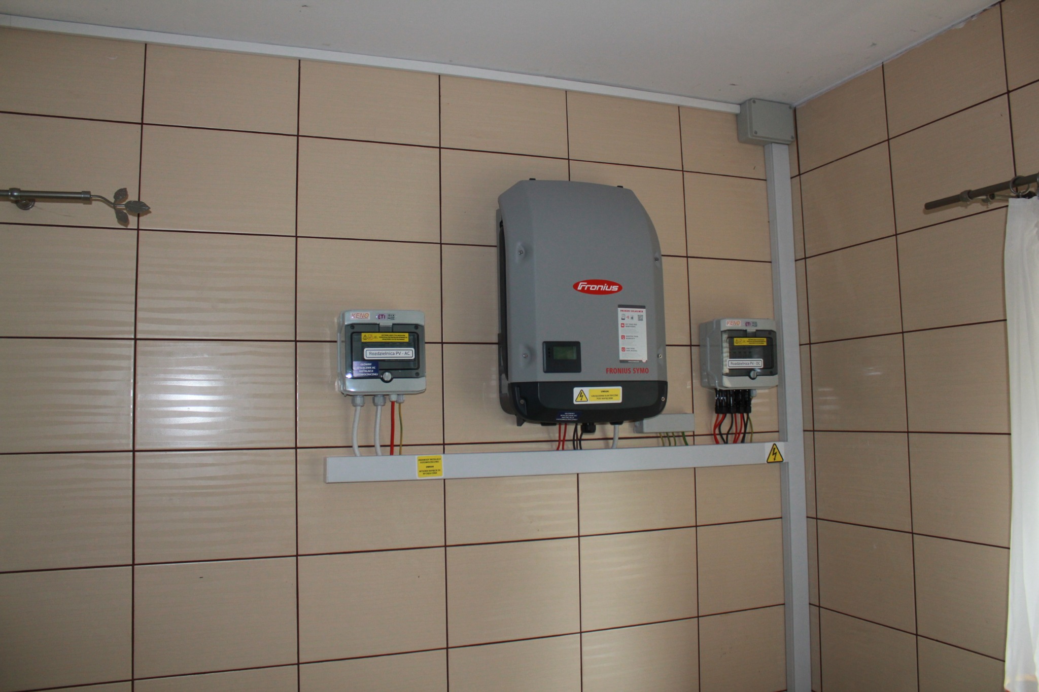 Szary falownik Fronius Symo zamontowany na ścianie wyłożonej jasnymi, kwadratowymi płytkami ceramicznymi, obok dwie szare rozdzielnice PV-AC, widoczne kable i żółte naklejki ostrzegawcze.