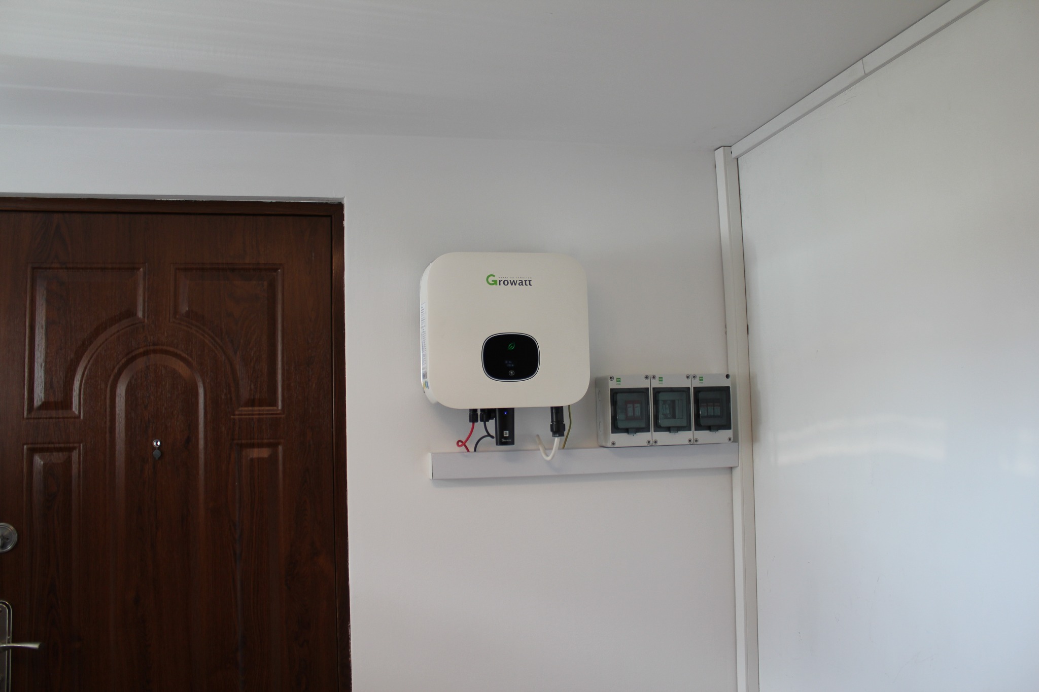 Biały inwerter Growatt zamontowany na ścianie wewnątrz budynku, obok brązowych drzwi wejściowych i skrzynki z bezpiecznikami.