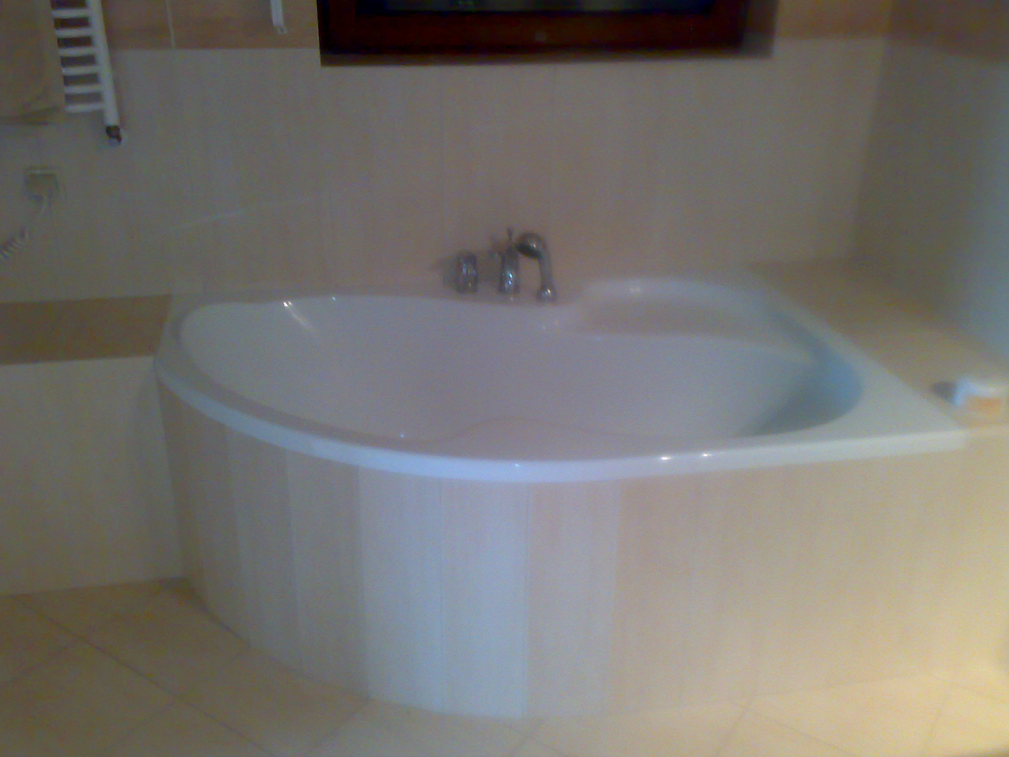 Biała, narożna wanna z chromowaną armaturą w wykończonej łazience z beżowymi płytkami i grzejnikiem drabinkowym.