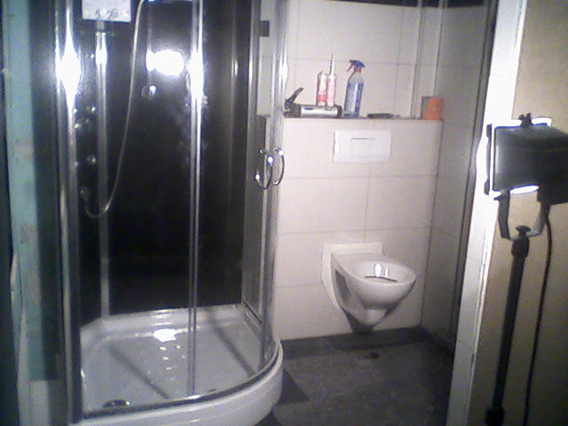 Wnętrze łazienki w trakcie remontu: kabina prysznicowa z hydromasażem, wisząca toaleta i akcesoria na półce, oświetlenie studyjne po prawej.