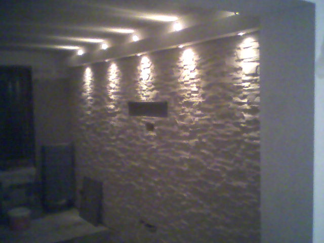 Wykończona ściana z dekoracyjnej imitacji kamienia podświetlona punktowymi lampami sufitowymi, widoczny fragment sufitu i narożnika ściany, na podłodze wiaderko i płyta G/K.