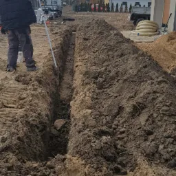 Wąski wykop w ziemi, obok pracownik z niwelatorem, w tle zabudowa jednorodzinna i zaparkowany samochód dostawczy z reklamą firmy.