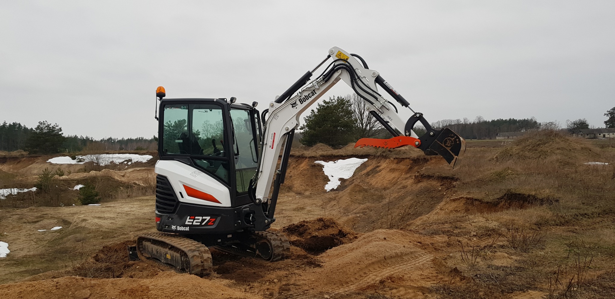 Kompaktowa koparka Bobcat E27z z pomarańczowym zębem chwytaka, pracująca na piaszczystym terenie z resztkami śniegu w tle, pochmurne niebo.