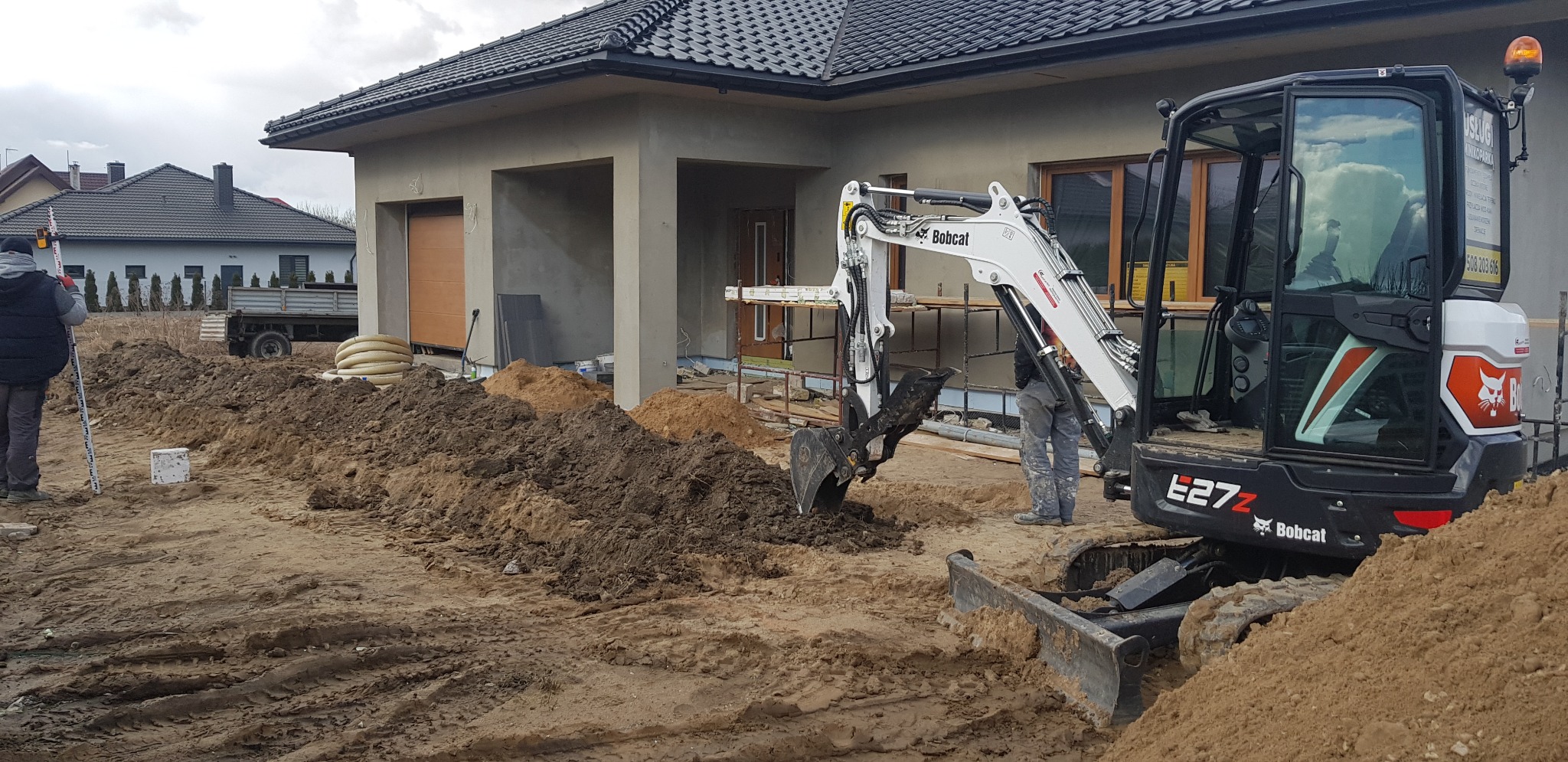Biała mini koparka Bobcat E27Z pracuje przy wykopie obok domu w trakcie budowy; widoczny pracownik z niwelatorem optycznym.