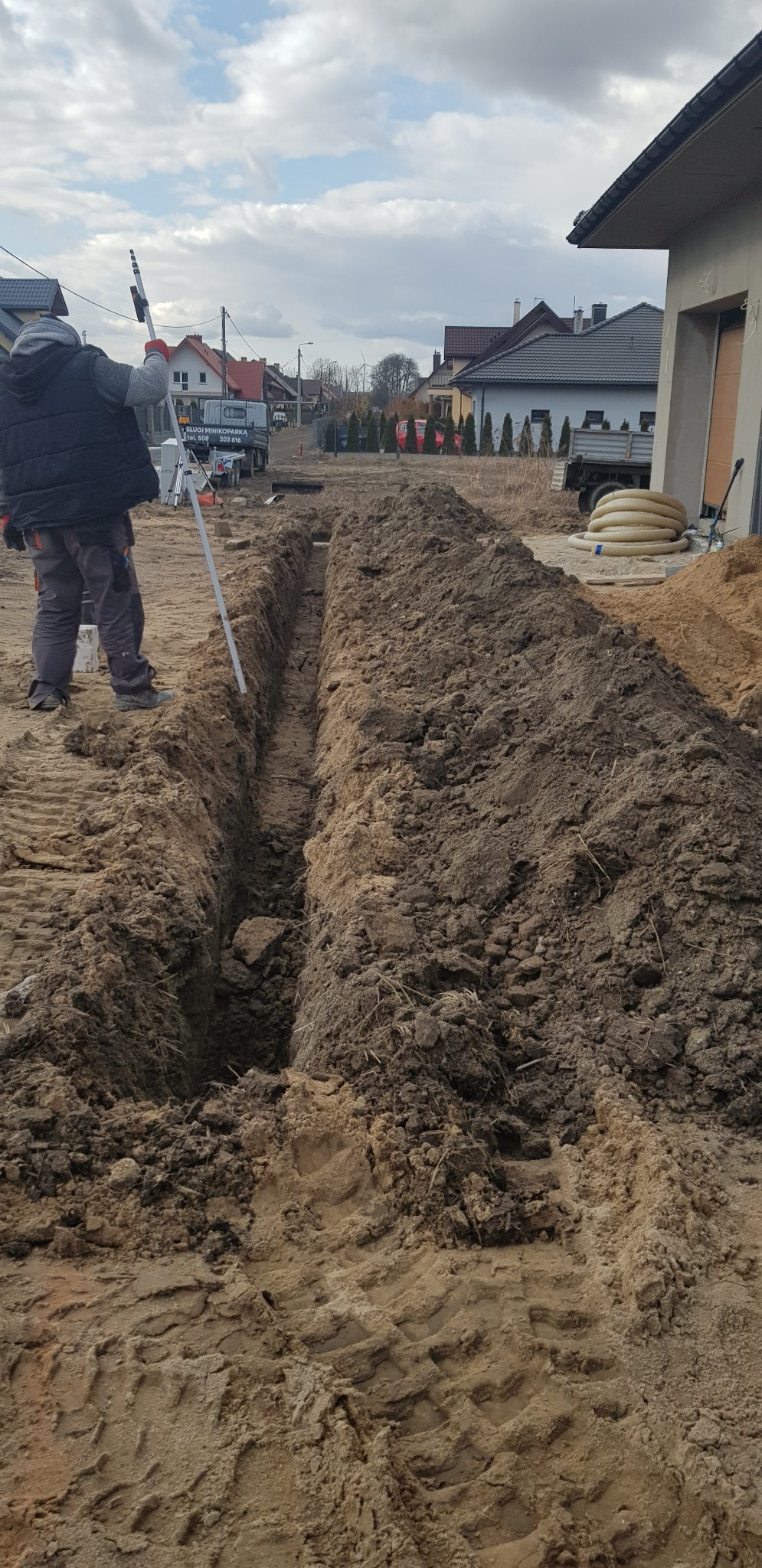 Wąski wykop w ziemi, obok pracownik z niwelatorem, w tle zabudowa jednorodzinna i zaparkowany samochód dostawczy z reklamą firmy.