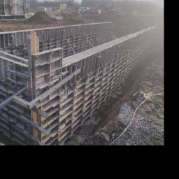 Szare szalunki systemowe tworzące formę pod wylanie betonu na mury oporowe, widoczne zbrojenia i grunt, w tle budowa i koparka.