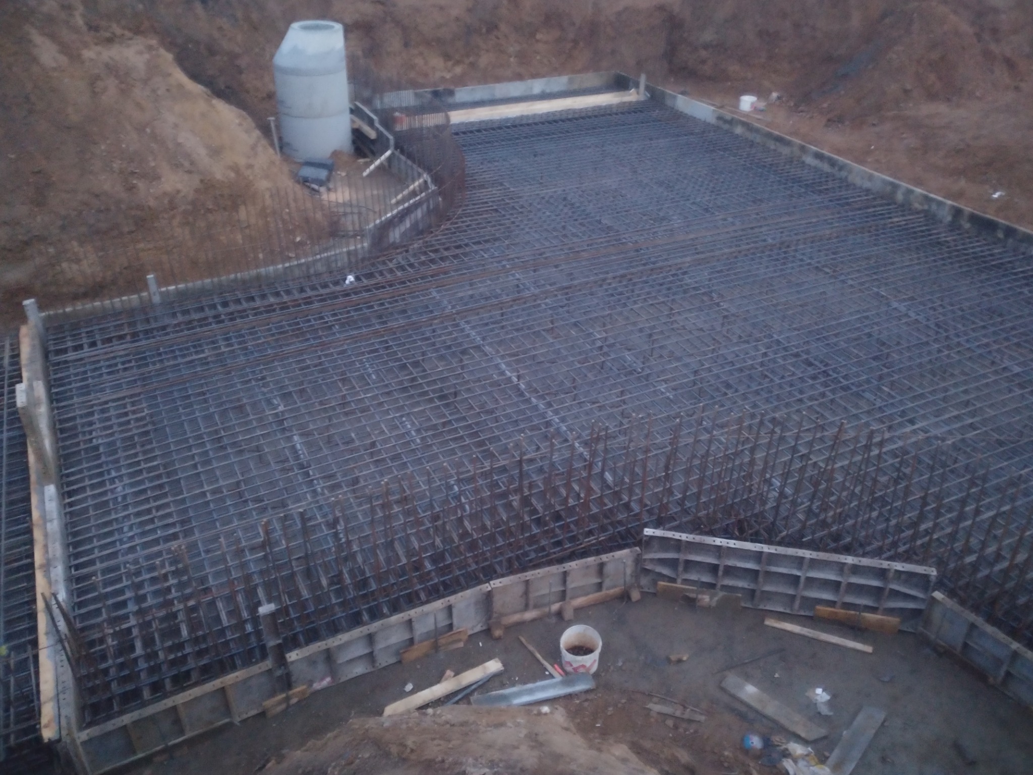 Siatka zbrojeniowa ułożona w wykopie, z widocznymi szalunkami i rurą betonową w tle, przygotowana do zalania betonem.