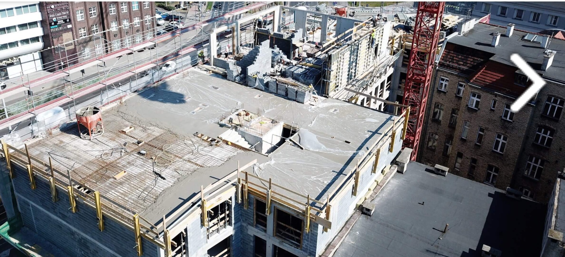 Wysoka perspektywa na plac budowy z widoczną konstrukcją żelbetową, zalewanym betonem stropem, zbrojeniami i czerwonym dźwigiem w tle miejskiej zabudowy.