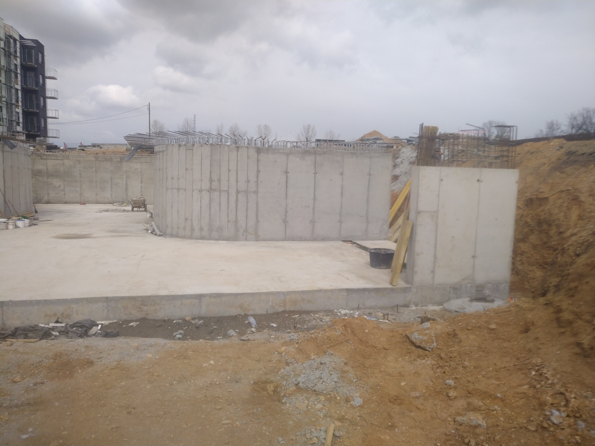 Wykop pod budynek z gotowymi betonowymi ścianami fundamentowymi, widoczne zbrojenia i przygotowany teren pod wylewkę, na tle pochmurnego nieba i powstającego budynku.