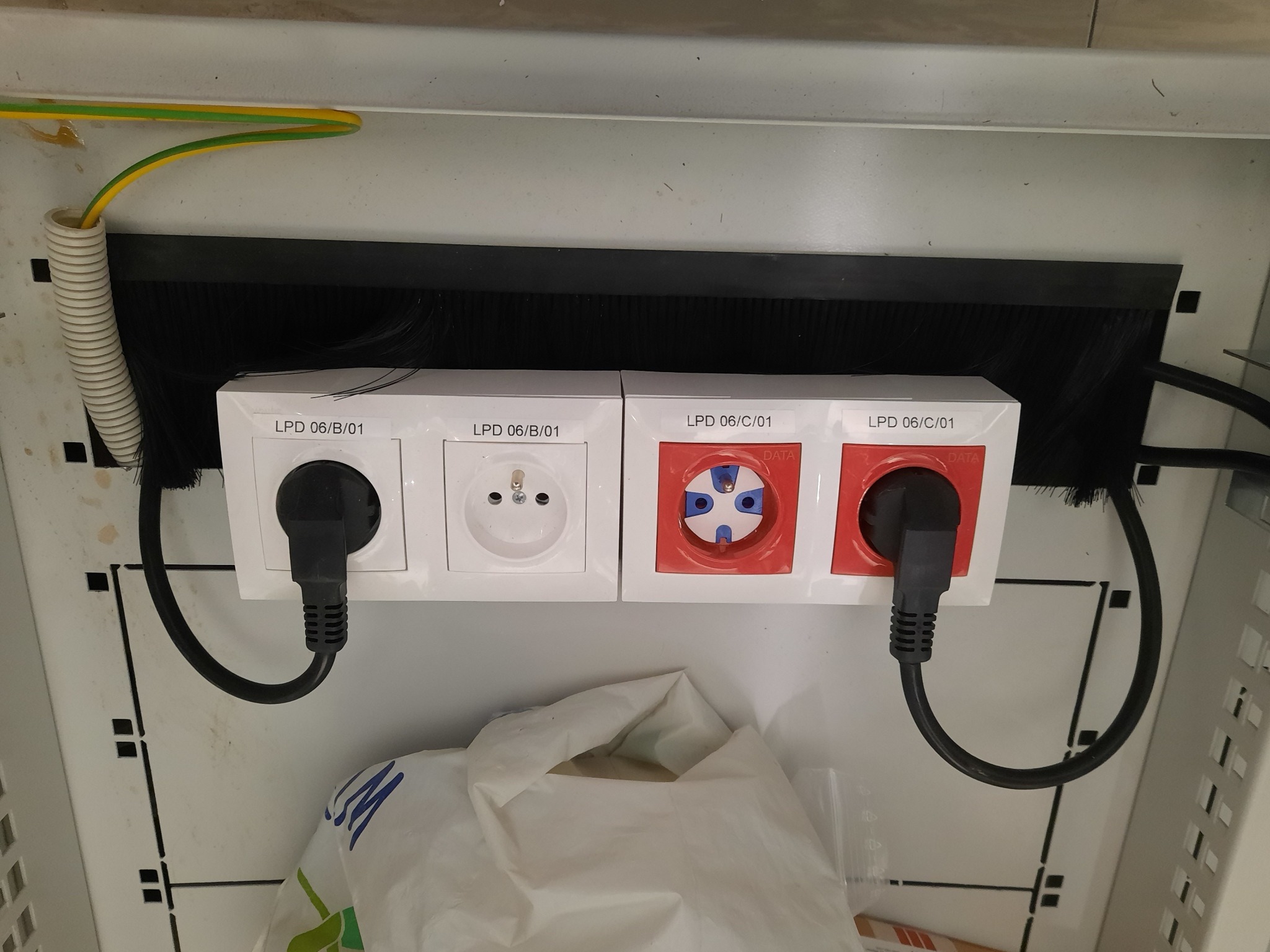 Panel z dwoma gniazdami elektrycznymi (europejskim i przemysłowym) zamontowany w szafie, z podłączonymi wtyczkami i widocznym przewodem ochronnym w peszlu.