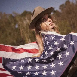 Portret kobiety w kapeluszu, owiniętej flagą USA na tle błękitnego nieba i zieleni. Artystyczne ujęcie z delikatnym filtrem, podkreślające patriotyczny charakter.