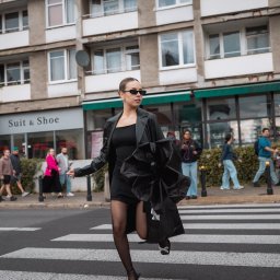 Aleksandra Żurek - Kobieta w czarnym stroju biegnie przez przejście dla pieszych na tle miejskiego krajobrazu z budynkami i przechodniami. Uwagę przyciągają okulary i nietypowa torebka.