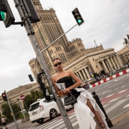 Aleksandra Żurek - Stylowa sesja zdjęciowa modelki w Warszawie na tle Pałacu Kultury, trzymającej gazetę i precla, oparta o słup sygnalizacji świetlnej na przejściu dla pieszych.