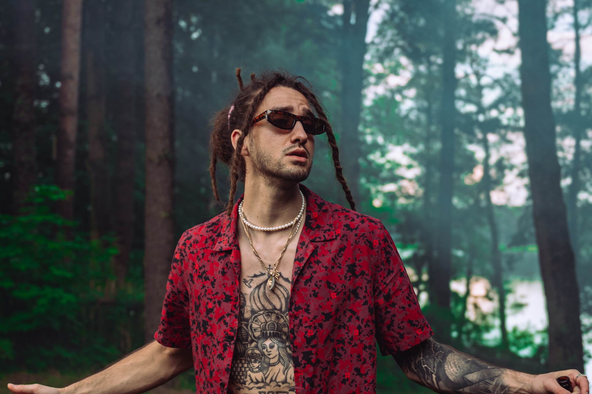 Mężczyzna z dredami w czerwonej koszuli, okularach i tatuażami na tle zamazanego lasu. Nietypowa stylizacja i artystyczny wyraz.