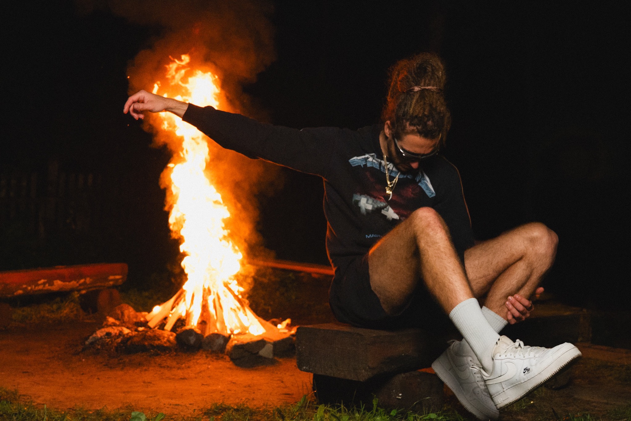 Mężczyzna z dredami siedzi blisko ogniska w nocy, ubrany w czarną bluzę i białe buty sportowe, z rozpostartą ręką w stronę płomieni. Nietypowe ujęcie.