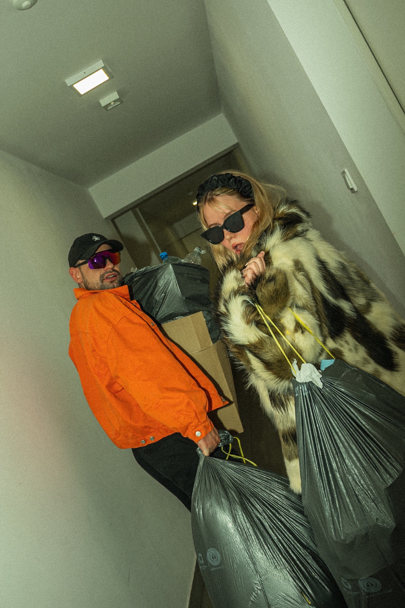 Ekstrawagancka para w okularach przeciwsłonecznych niesie worki ze śmieciami w korytarzu. Mężczyzna w pomarańczowej kurtce, kobieta w futrze. Niska perspektywa.