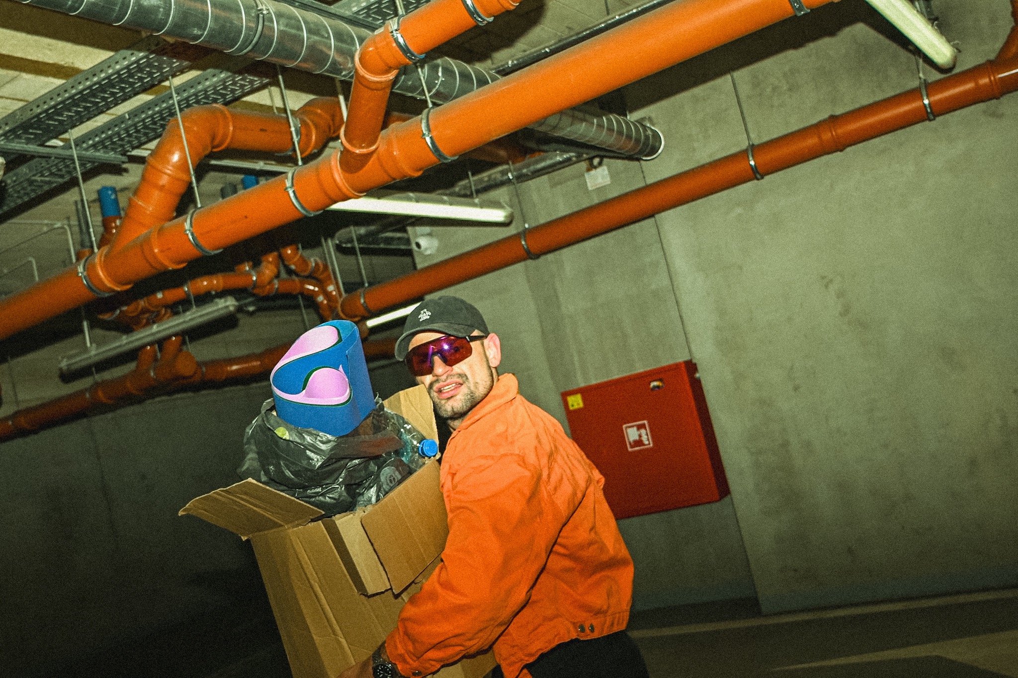 Mężczyzna w pomarańczowej kurtce niesie karton z odpadami na tle instalacji rurowej i ściany. Nietypowe ujęcie w przestrzeni technicznej.