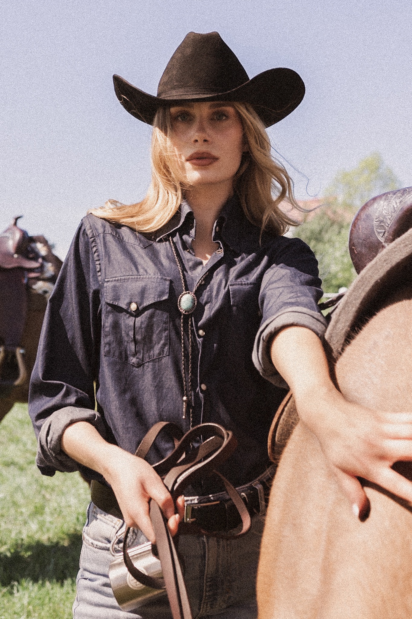 Kobieta w kapeluszu kowbojskim, dżinsowej koszuli i bolo tie trzyma lejce, opierając się o konia. W tle łąka i drugi koń z siodłem.