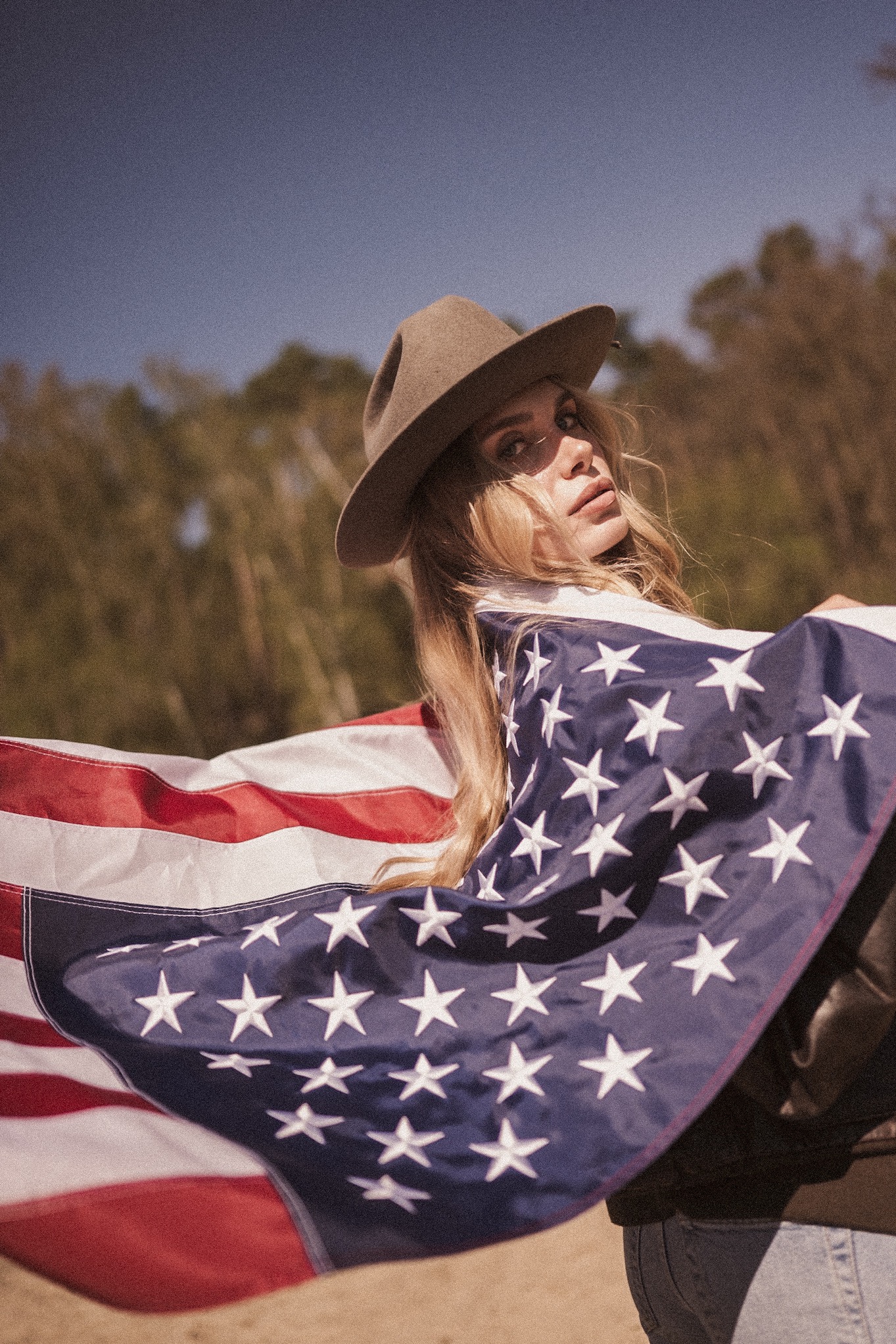 Portret kobiety w kapeluszu, owiniętej flagą USA na tle błękitnego nieba i zieleni. Artystyczne ujęcie z delikatnym filtrem, podkreślające patriotyczny charakter.