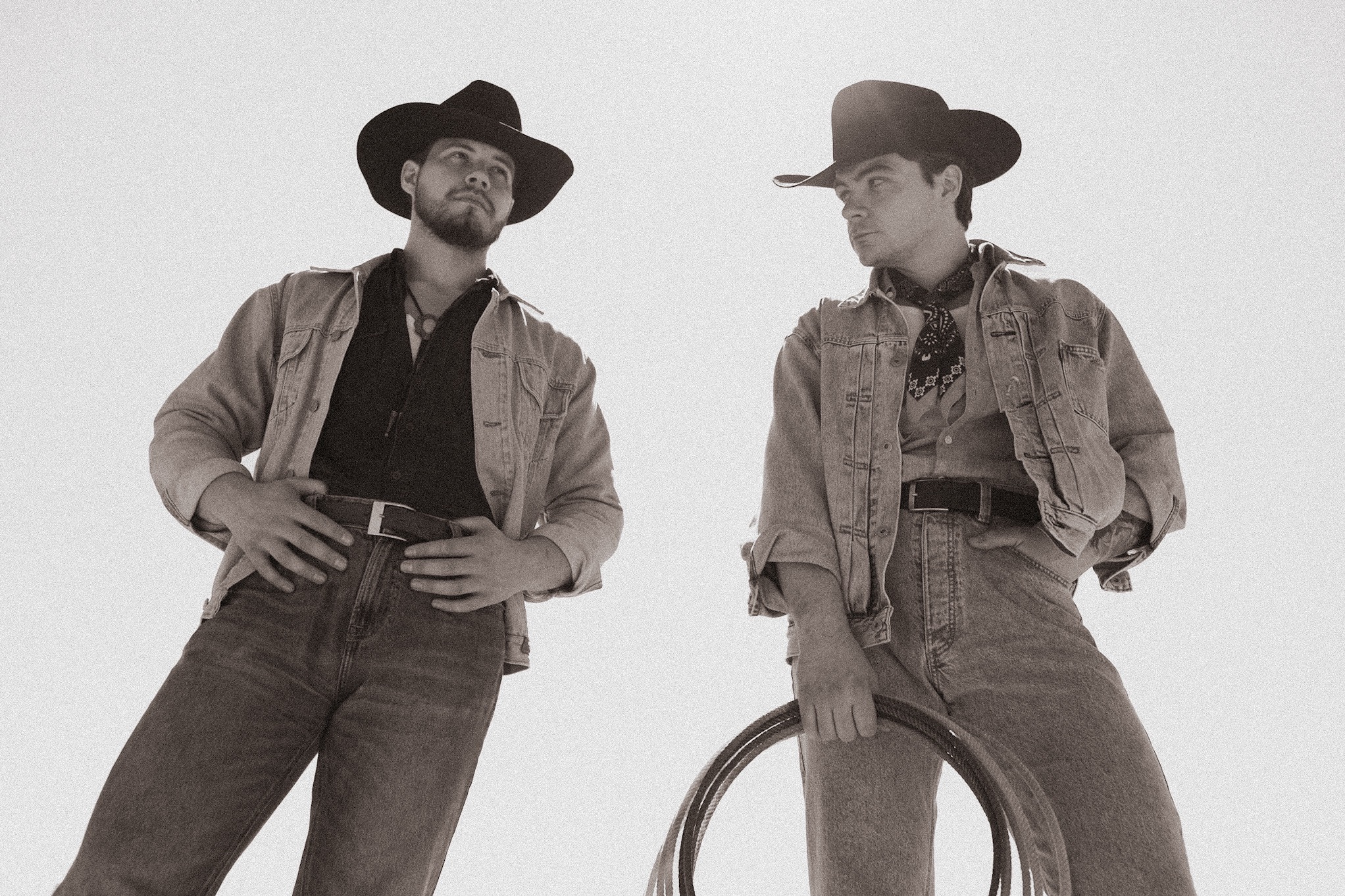 Dwóch mężczyzn w kowbojskich kapeluszach i dżinsowych kurtkach, niski kąt ujęcia, stylizowane na czarno-białe, jeden trzyma lasso, spoglądają na siebie.