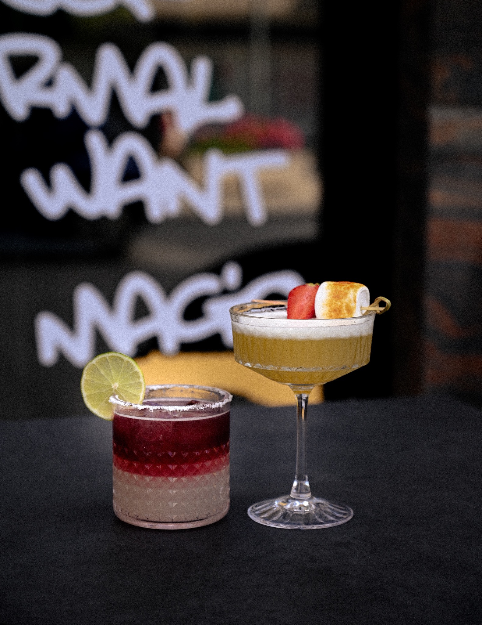 Dwa koktajle: bordowy z limonką i złoty z pianką i truskawką, na tle napisu na szybie, w barowych klimatach.