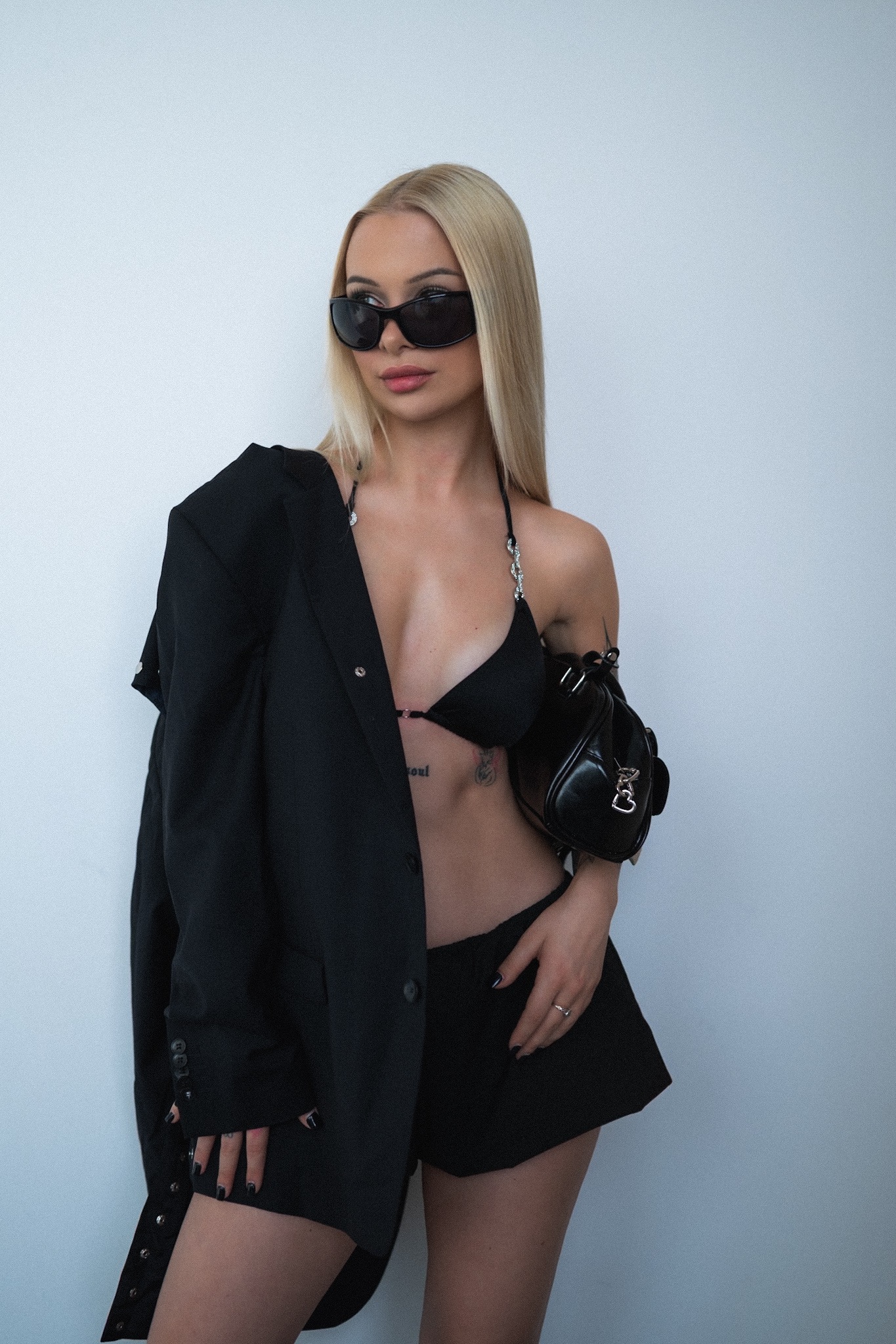 Blondwłosa kobieta w czarnym bikini i marynarce, z torebką, pozuje na tle białej ściany. Uwagę zwracają czarne okulary i tatuaż na brzuchu.