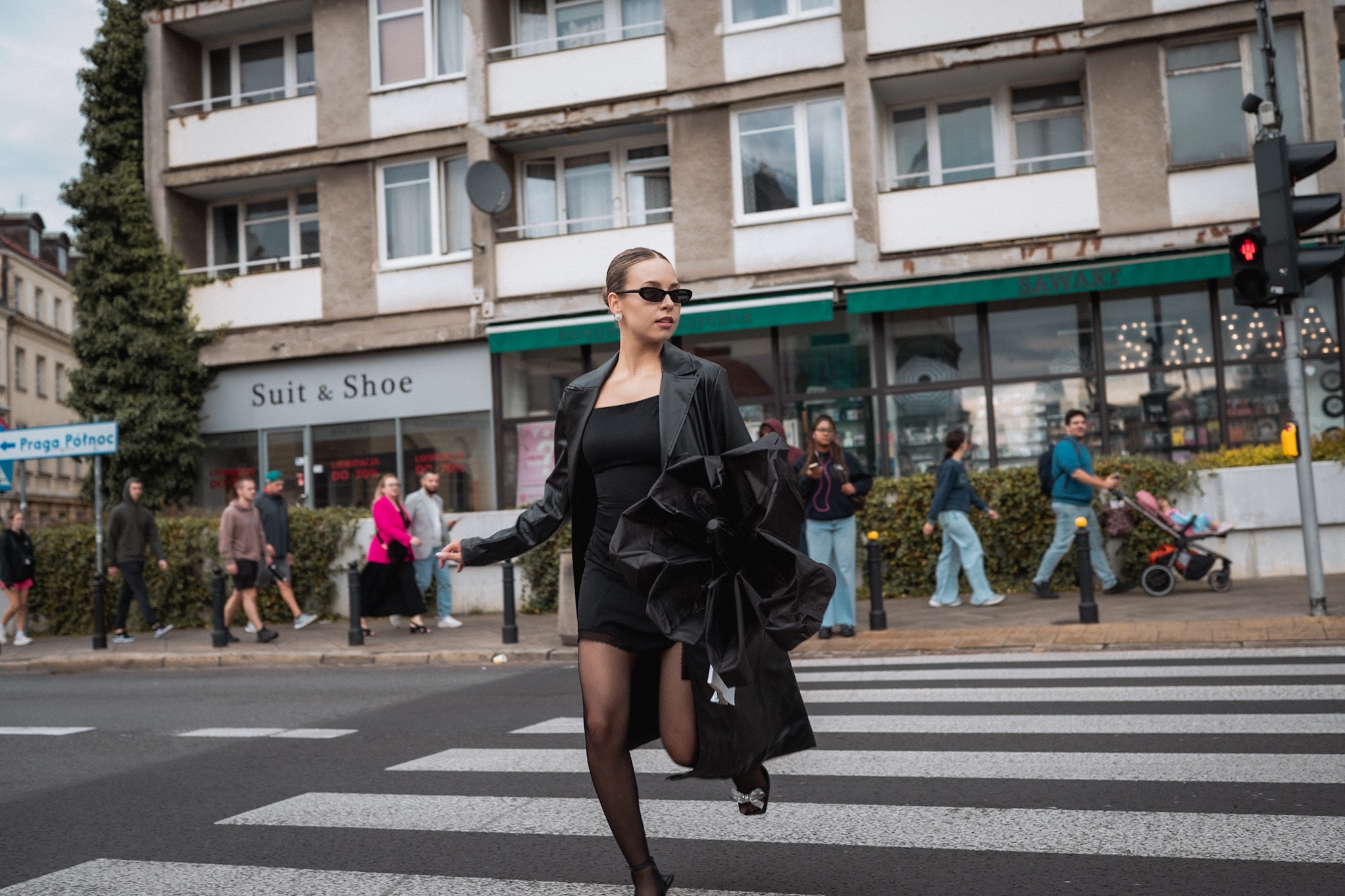 Kobieta w czarnym stroju biegnie przez przejście dla pieszych na tle miejskiego krajobrazu z budynkami i przechodniami. Uwagę przyciągają okulary i nietypowa torebka.