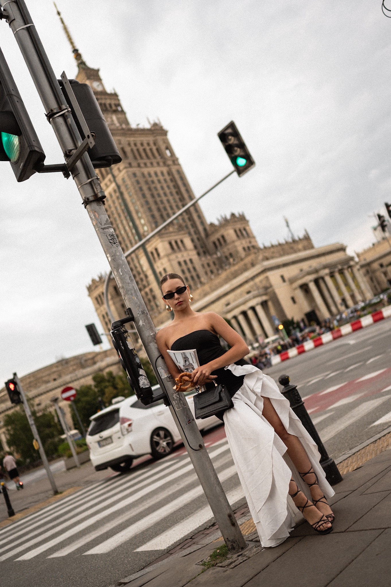 Stylowa sesja zdjęciowa modelki w Warszawie na tle Pałacu Kultury, trzymającej gazetę i precla, oparta o słup sygnalizacji świetlnej na przejściu dla pieszych.