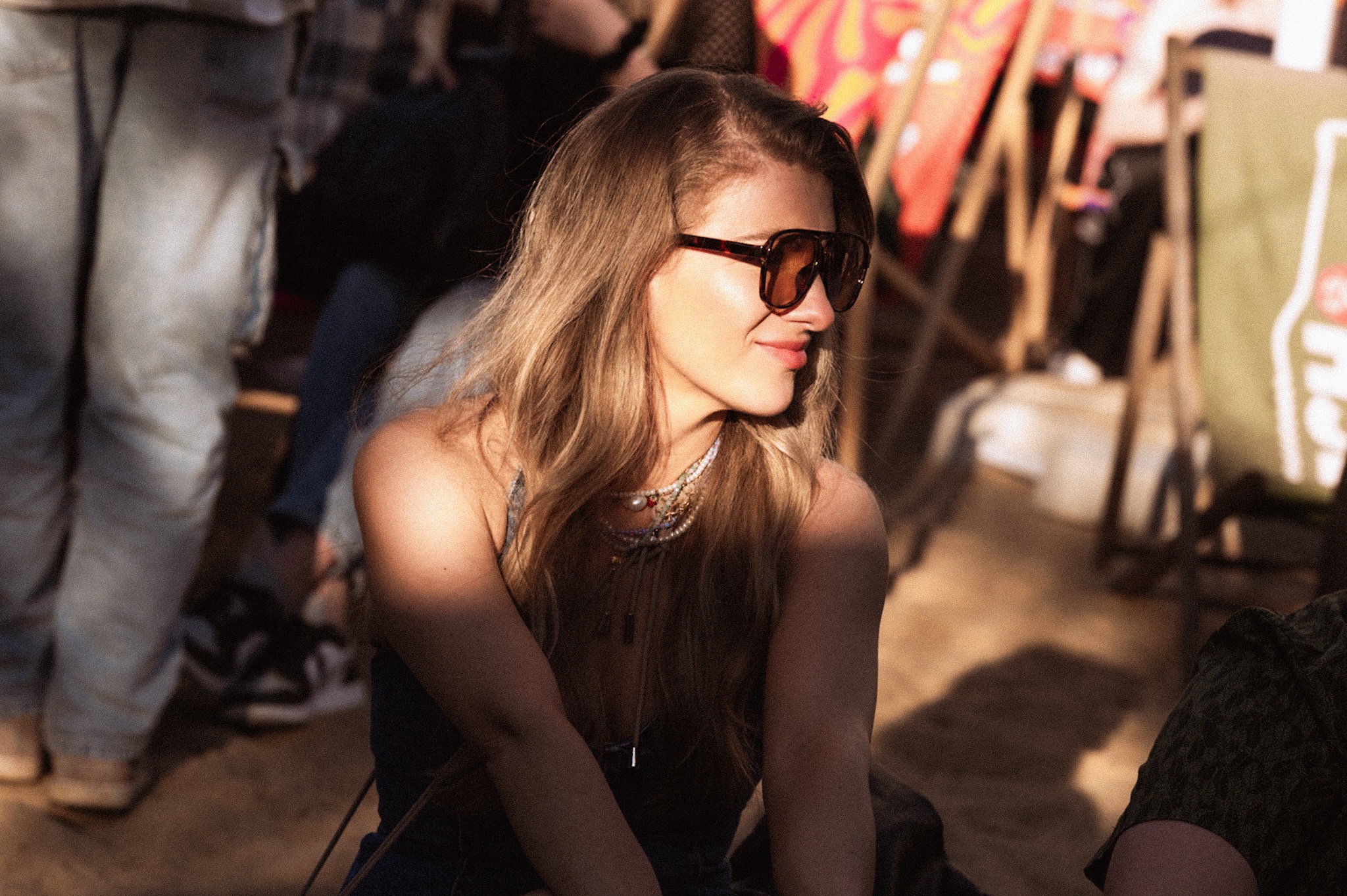 Portret kobiety w okularach przeciwsłonecznych, z delikatnym uśmiechem, w plenerze, z elementami plażowego otoczenia w tle, w ciepłym oświetleniu.