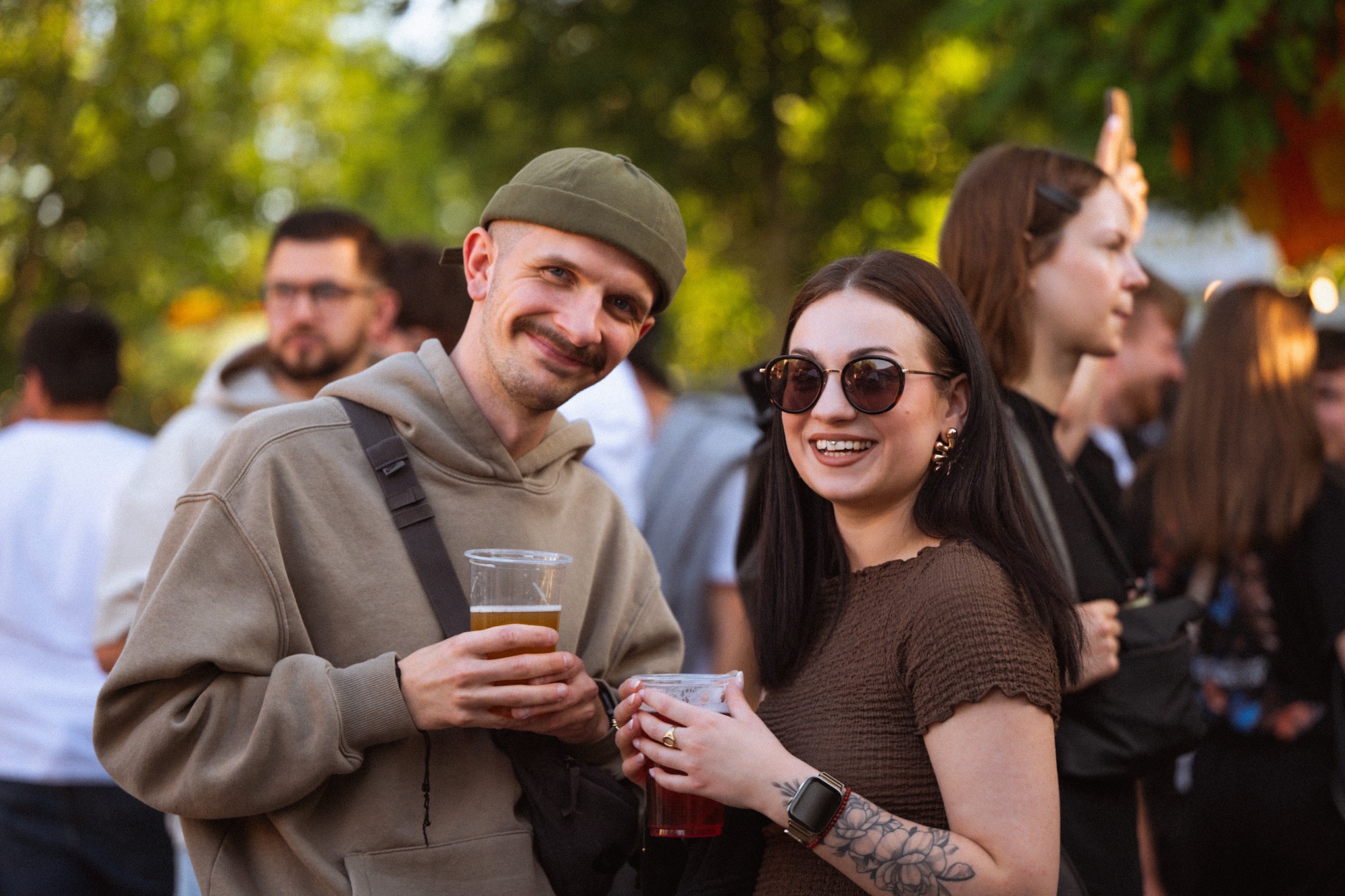 Uśmiechnięta para z napojami na tle rozmytej grupy ludzi i zieleni. Mężczyzna w czapce i bluzie, kobieta w okularach i z tatuażem. Atmosfera relaksu i zabawy.