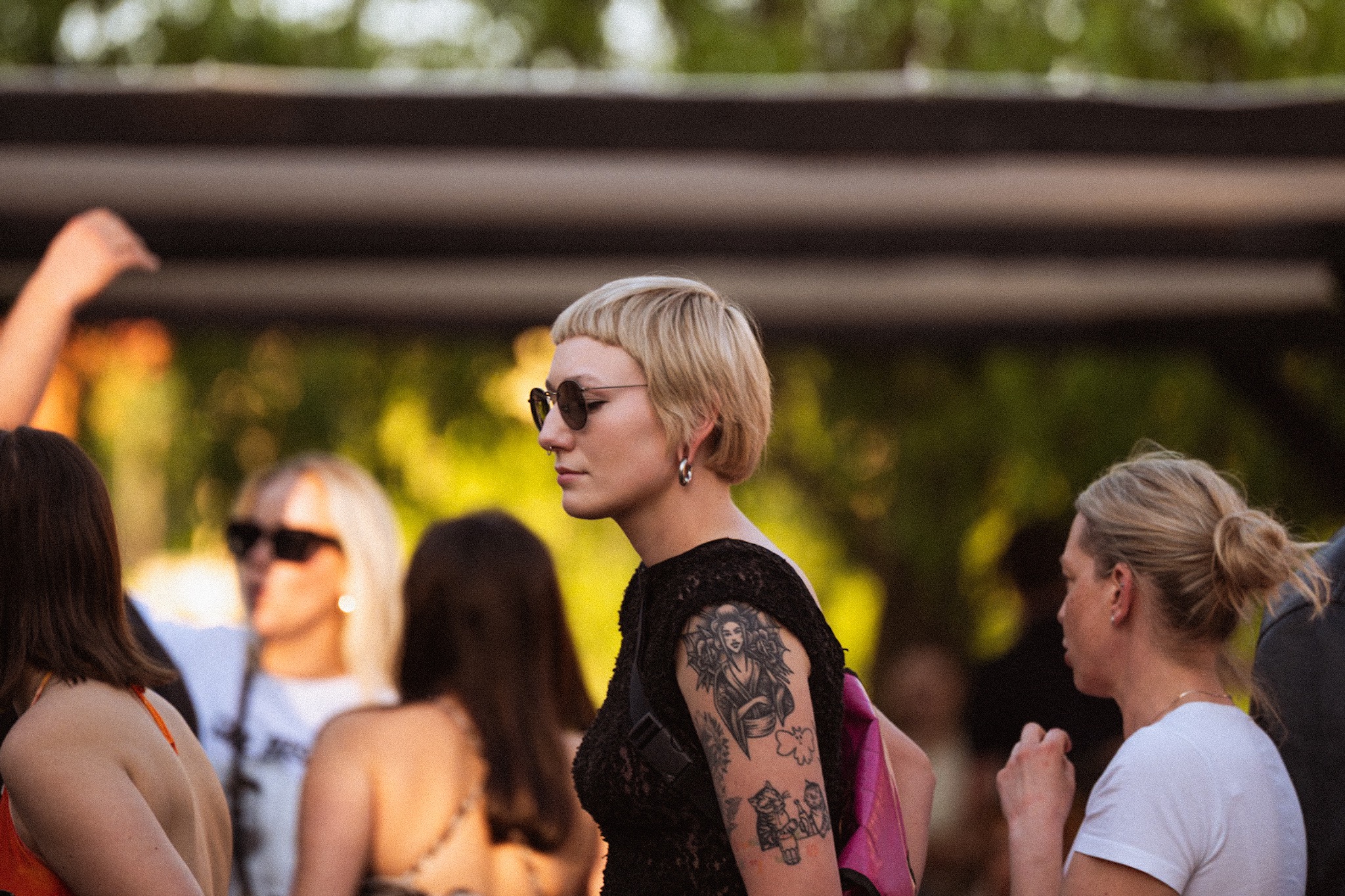 Kobieta z tatuażami na ramieniu, blond włosami i okularami przeciwsłonecznymi, w tłumie, z zarośniętym tłem.