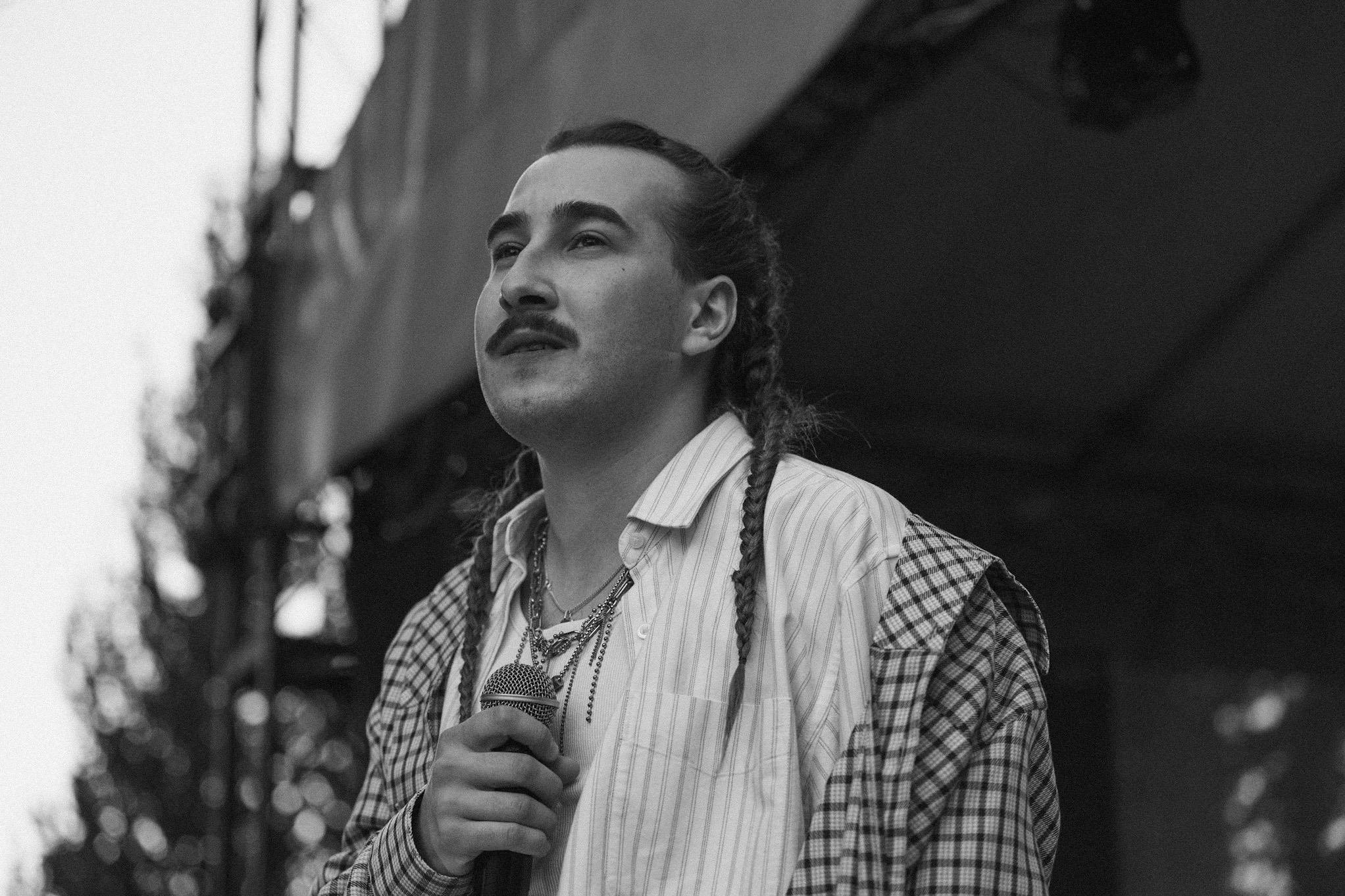 Czarno-białe zdjęcie mężczyzny z wąsami i warkoczami, trzymającego mikrofon, ubranego w koszulę w paski i kraciastą kurtkę, spoglądającego w górę.