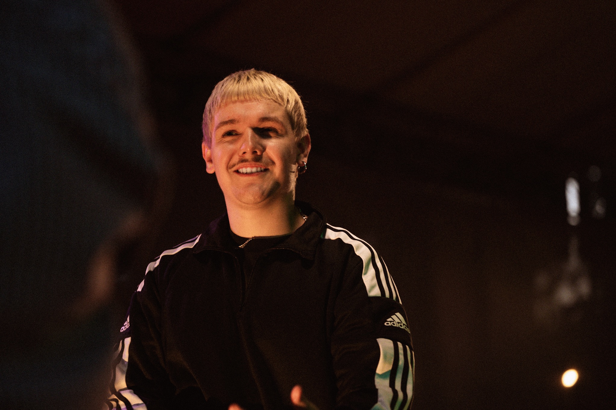 Uśmiechnięty mężczyzna z blond włosami i wąsami w czarnej bluzie Adidas z trzema paskami, stojący na tle ciemnej sceny oświetlonej punktowym światłem.