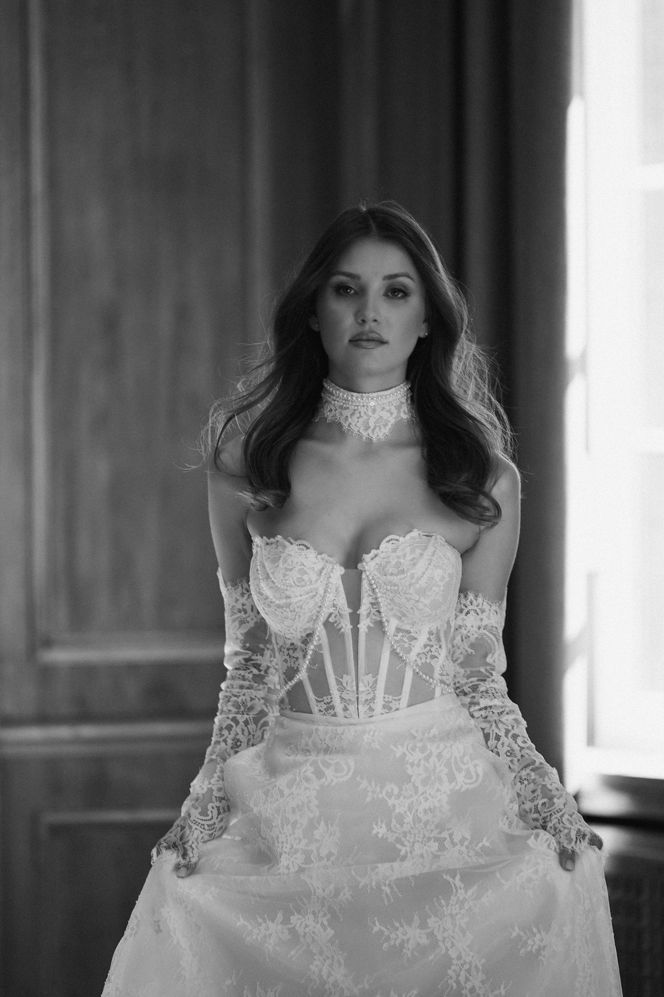 Czarno-białe zdjęcie kobiety w koronkowej sukni z chokerem, stojącej na tle ściany i okna, z delikatnym oświetleniem padającym zza niej.