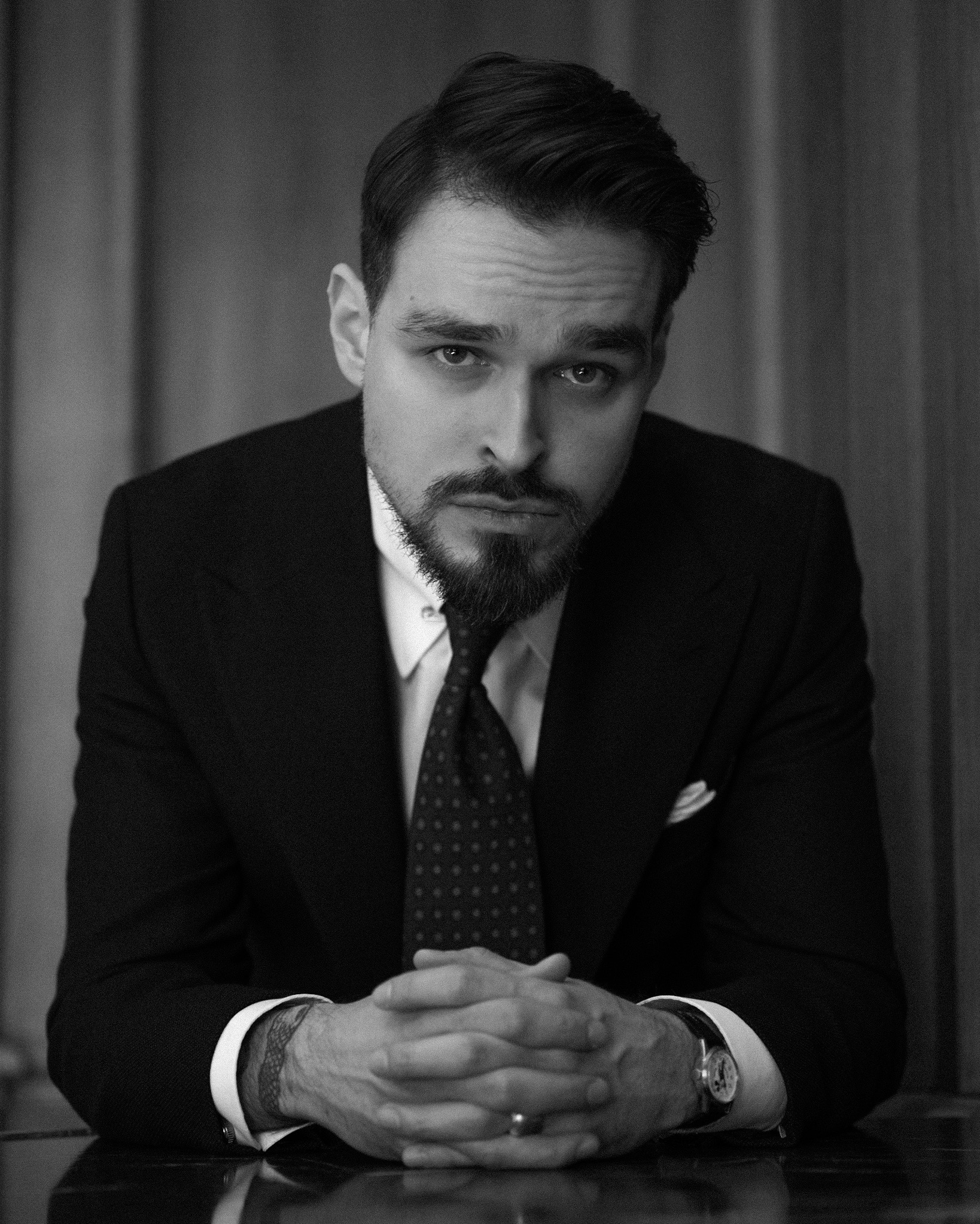 Portret biznesowy mężczyzny w garniturze, czarno-biały, z dłońmi splecionymi na blacie. Widoczny zegarek i tatuaż na nadgarstku, tło stanowią ciemne zasłony.