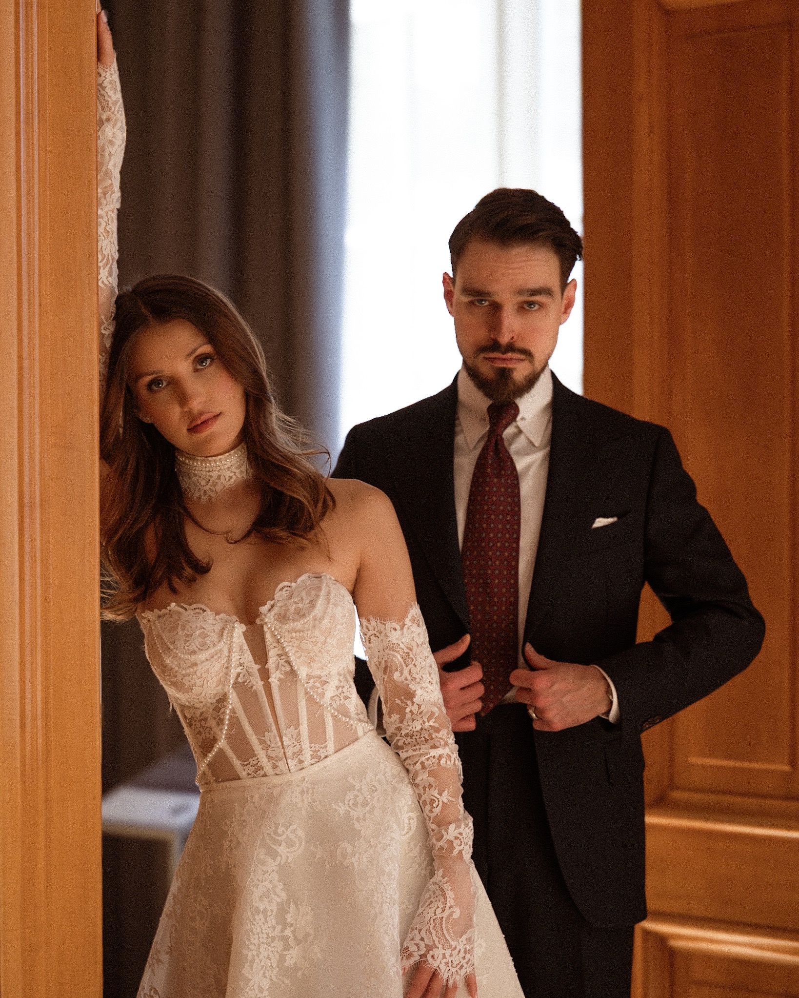 Elegancka sesja narzeczeńska: kobieta w koronkowej sukni i mężczyzna w garniturze, oboje z poważnym wyrazem twarzy, pozują w stylowym wnętrzu.