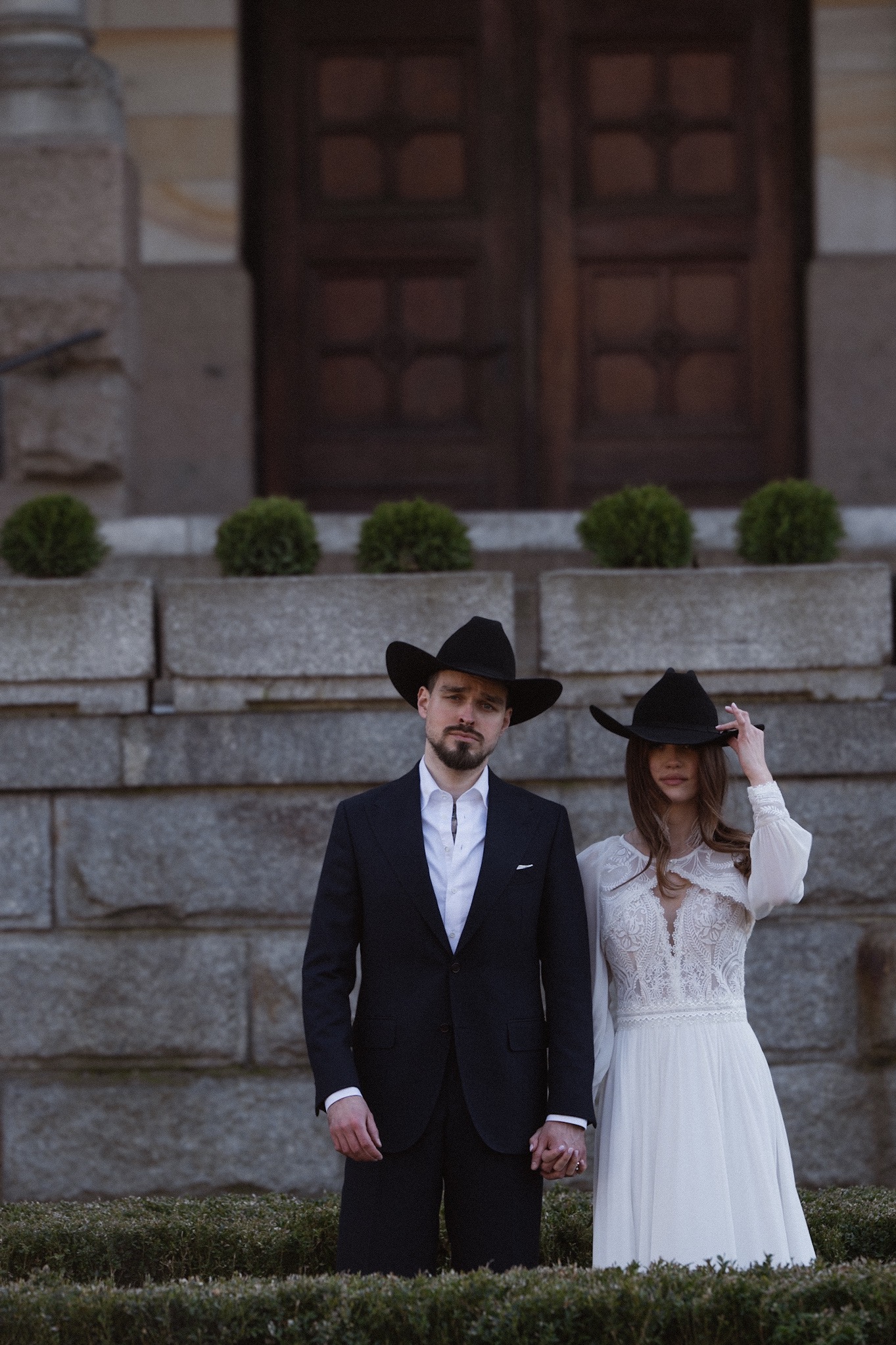 Para w strojach western na tle kamiennej architektury. On w czarnym garniturze, ona w białej sukni z koronką. Oboje w czarnych kapeluszach.