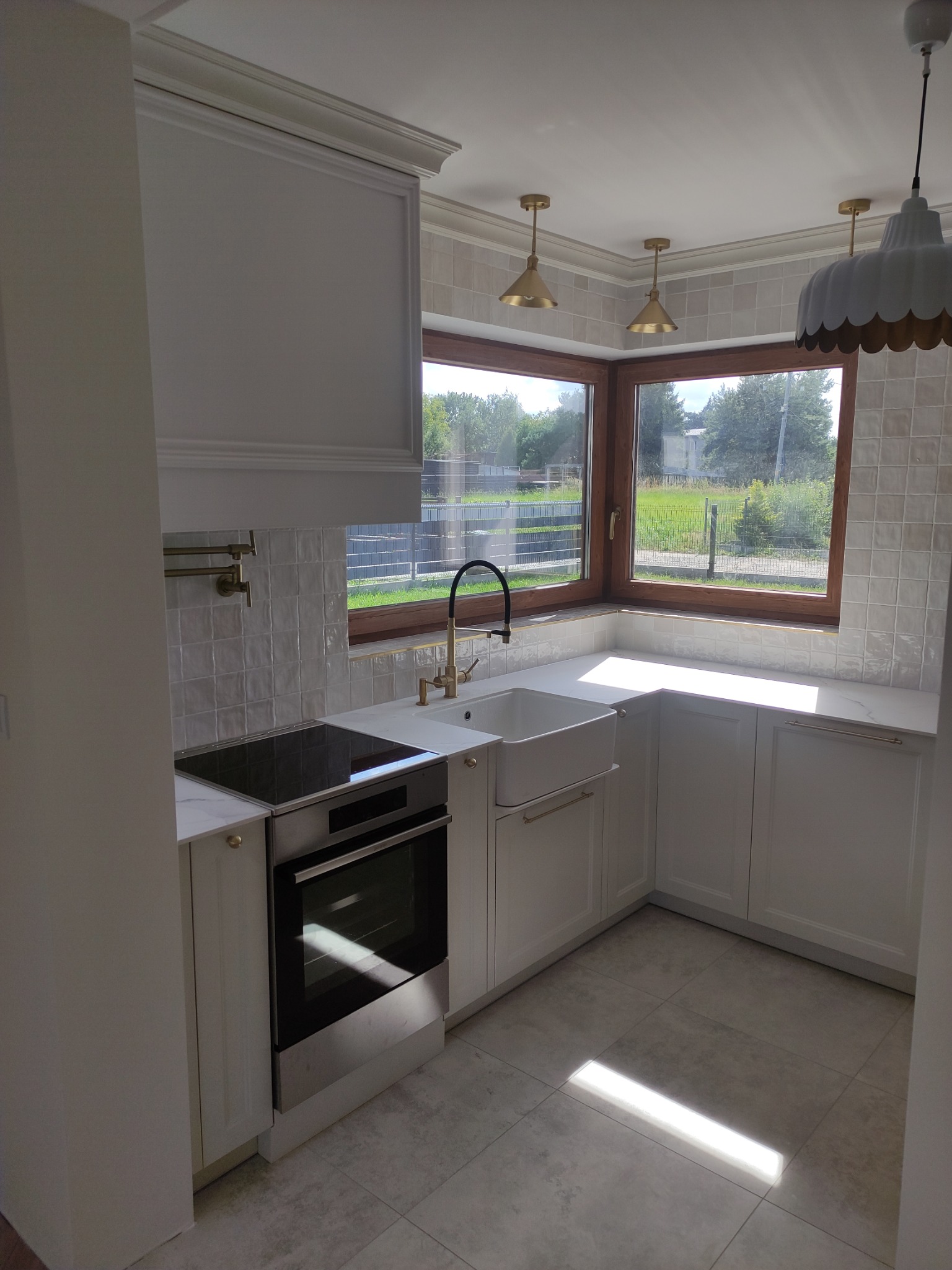 Jasna kuchnia w stylu klasycznym z białymi szafkami, ceramicznym zlewem farmerskim, złotą armaturą i płytkami ściennymi w odcieniach beżu, oświetlona trzema złotymi lampami i jedną białą lampą...