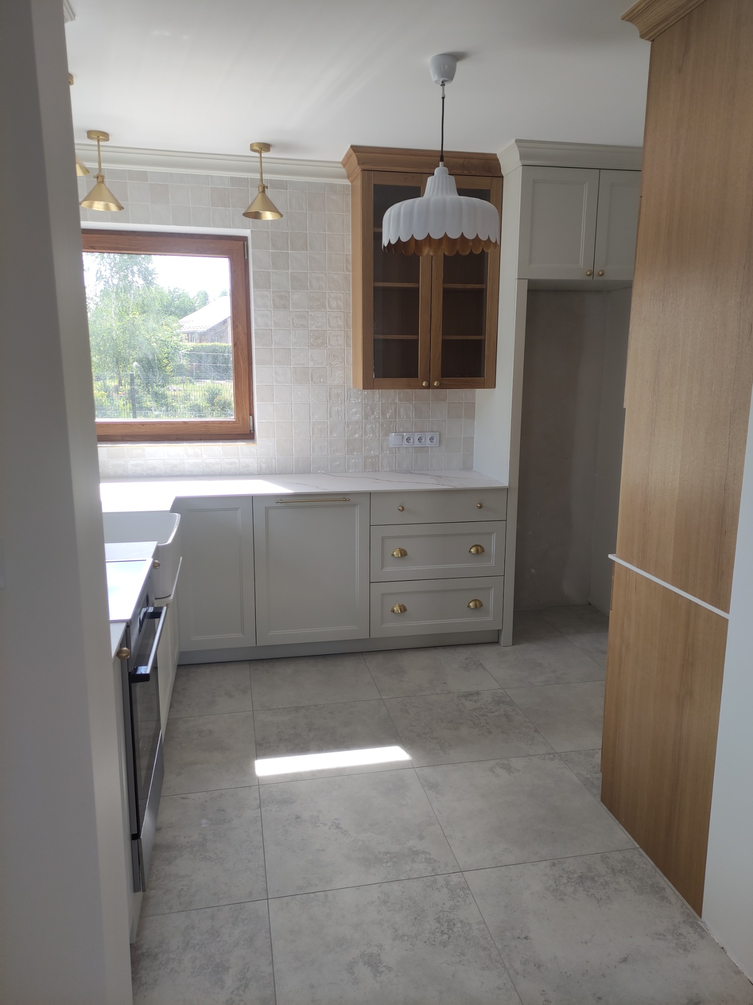 Jasna kuchnia z kremowymi szafkami z złotymi uchwytami, drewnianymi akcentami i płytkami na ścianie, oświetlona wiszącymi lampami.