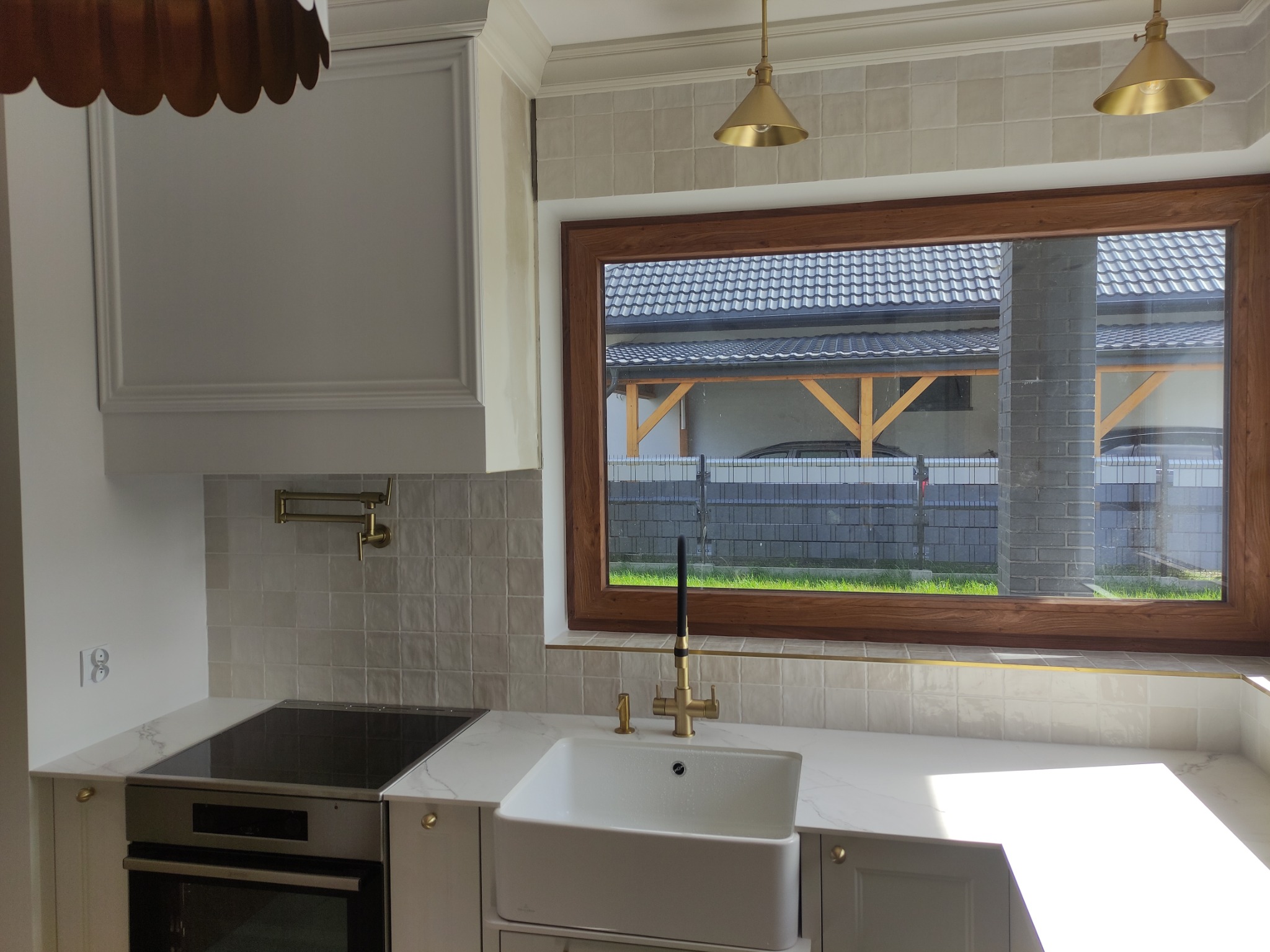 Nowoczesna kuchnia z białymi szafkami, blatem imitującym marmur, złotą armaturą i okładziną ścienną z kwadratowych płytek w odcieniach beżu. Przez okno widoczny ogród i zabudowania.