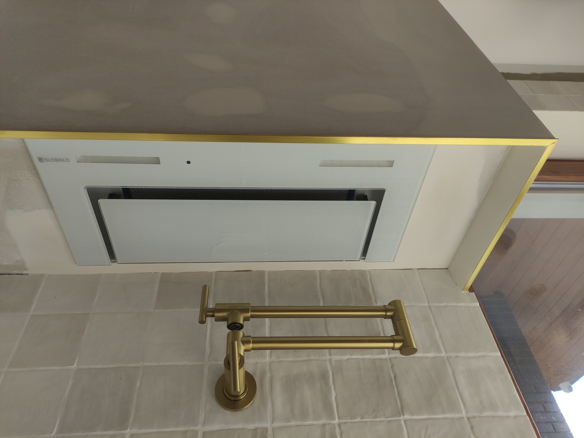 Nowoczesna kuchnia: biały okap z złotym wykończeniem nad płytą wyłożoną kwadratowymi, kremowymi płytkami i złotą baterią kuchenną z wysięgnikiem.