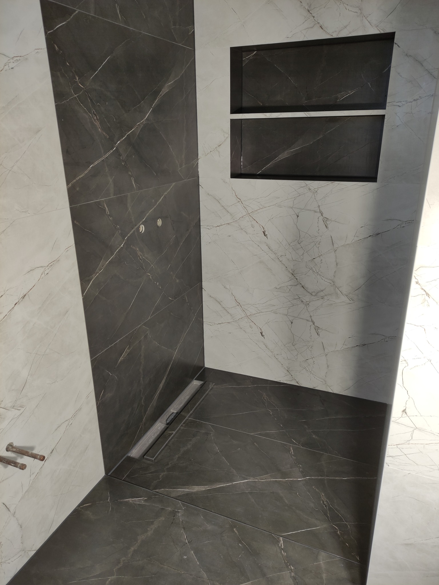 Nowoczesna kabina prysznicowa wyłożona płytkami imitującymi marmur w odcieniach szarości i bieli, z odpływem liniowym i wnęką z półkami.