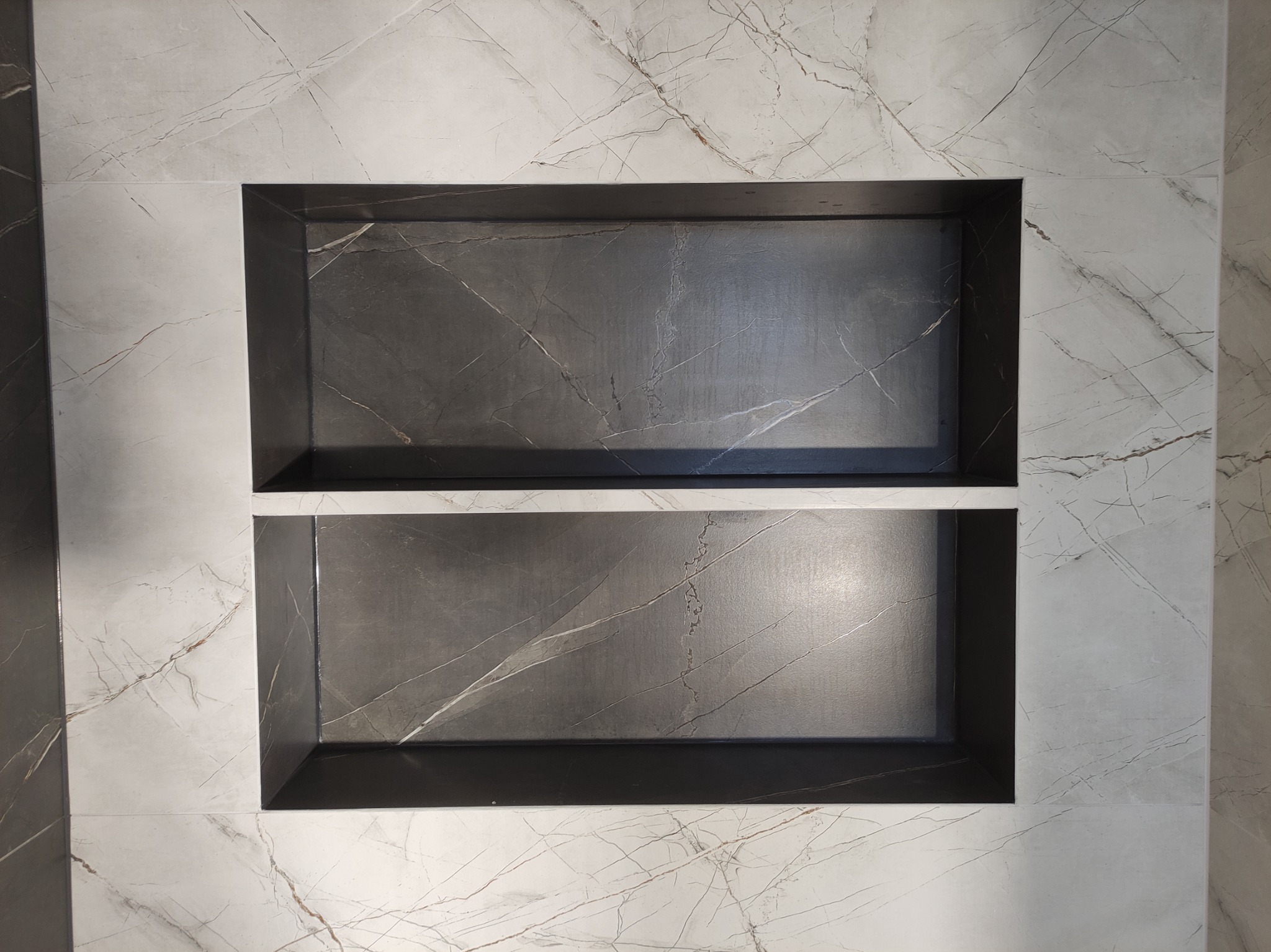 Dwie półki wnękowe wyłożone ciemnoszarymi płytkami z marmurkowym wzorem, otoczone jasnymi płytkami z podobnym wzorem żyłkowania.