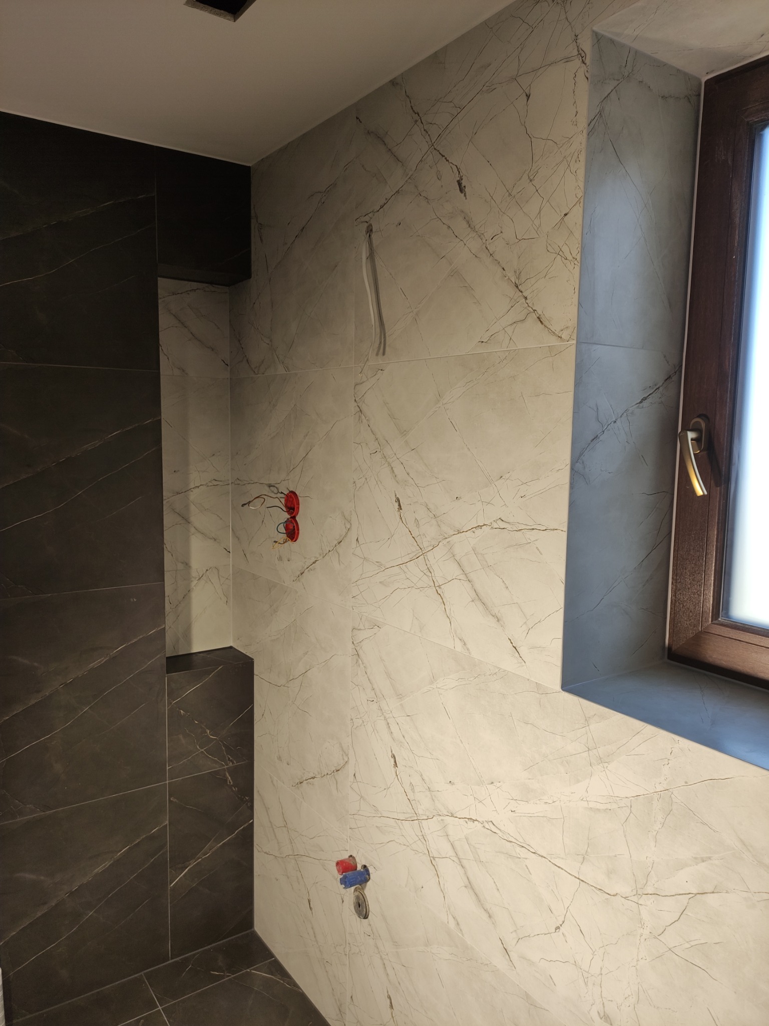 Ściany łazienki wyłożone płytkami imitującymi marmur w odcieniach szarości i bieli, widoczne wystające kable elektryczne i rury wodne, okno z brązową ramą.