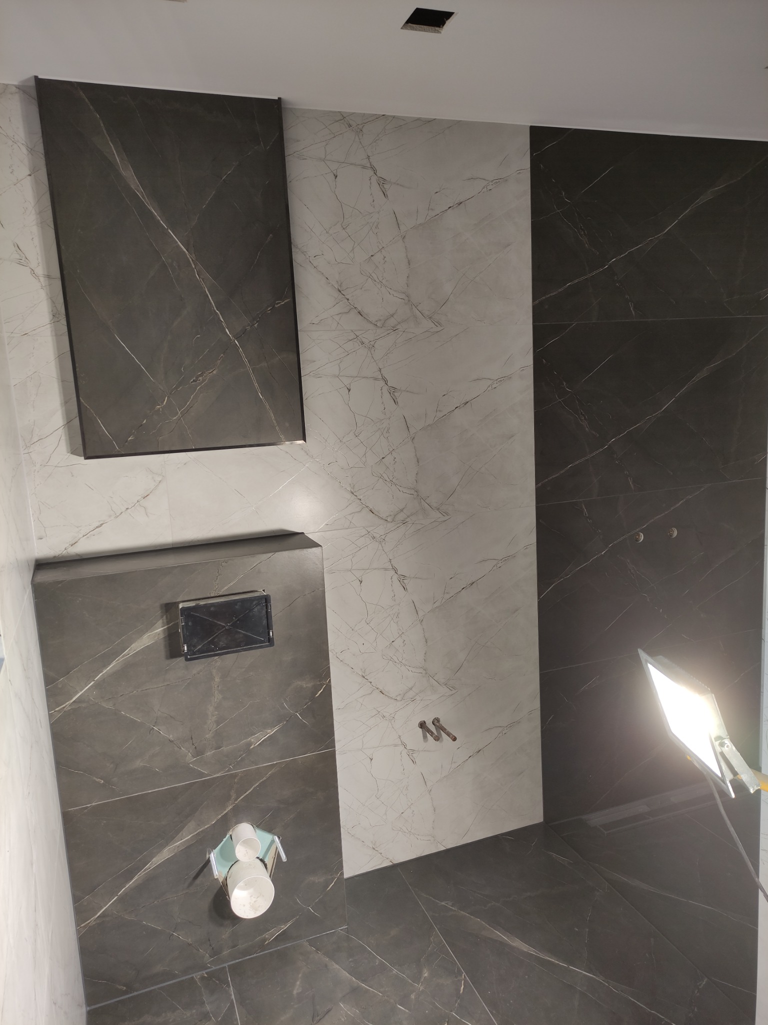 Częściowo wykończona łazienka z czarno-białymi płytkami imitującymi marmur, widoczne elementy instalacji wodnej i oświetlenie budowlane.