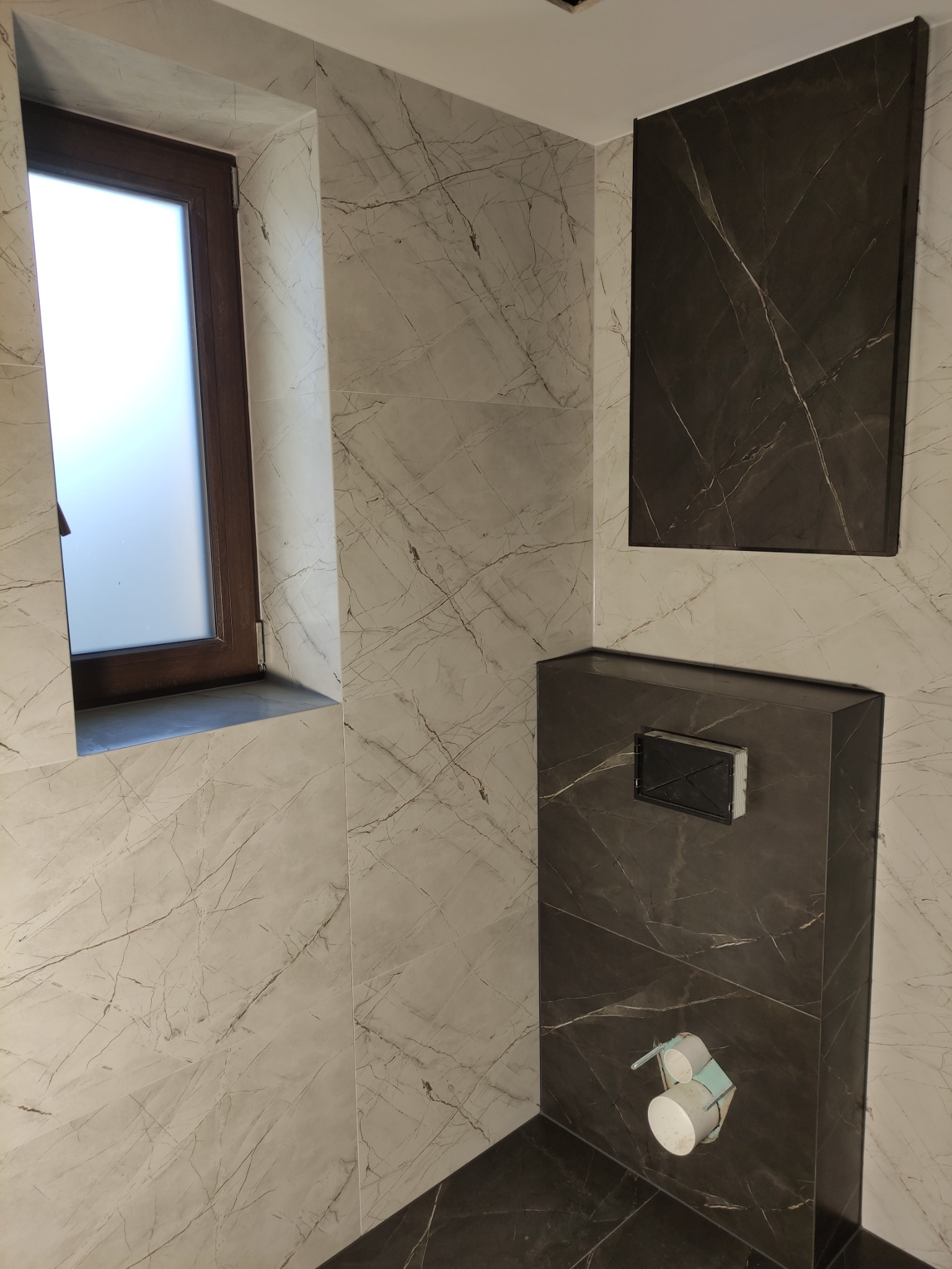 Wykończona łazienka z marmurowymi płytkami w odcieniach bieli i szarości, zabudowa WC z ciemnego marmuru, okno z drewnianą ramą, widoczny uchwyt na papier toaletowy.